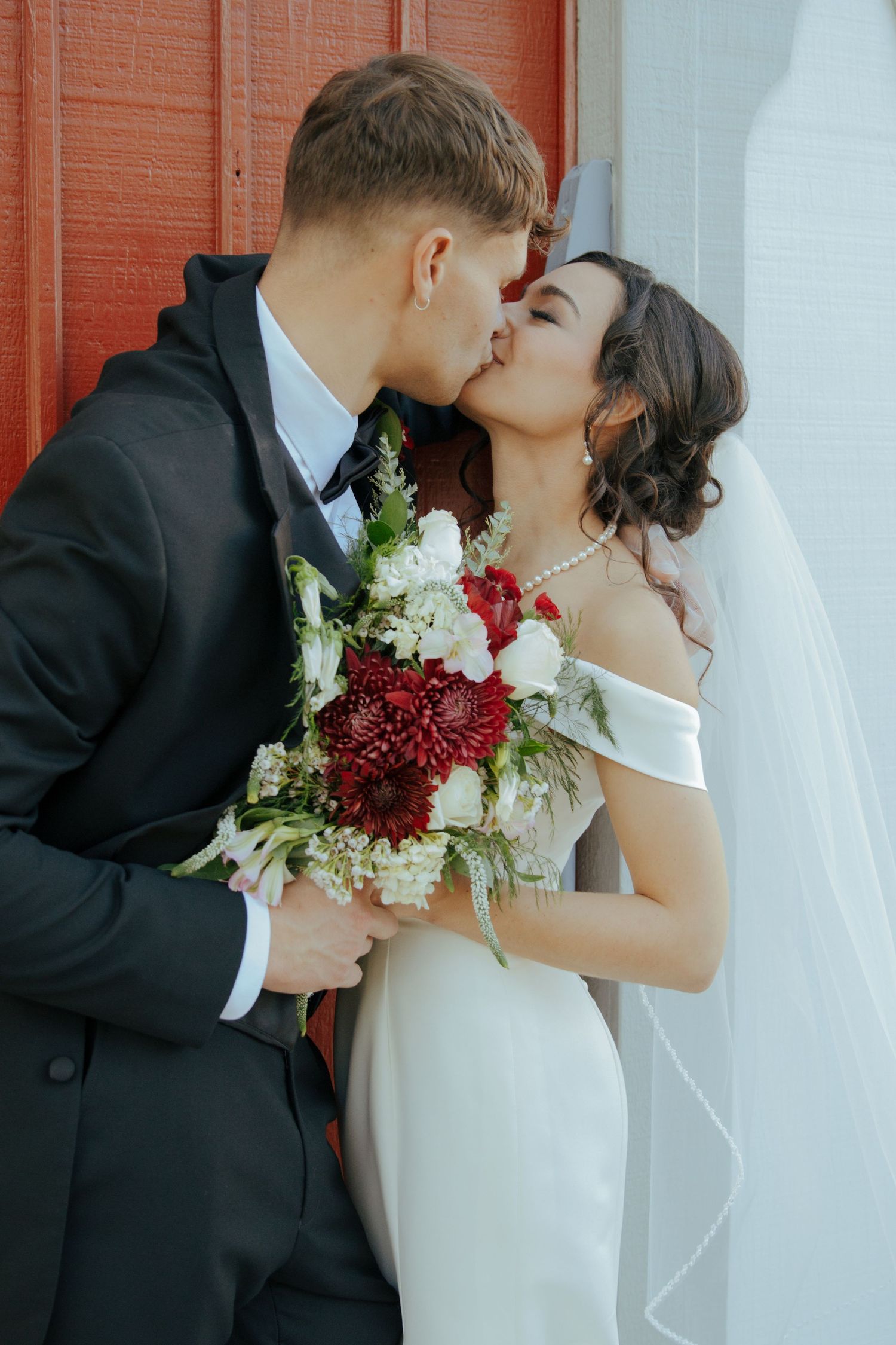 Bride and groom share kiss holding red and white floral bouquet against red door.