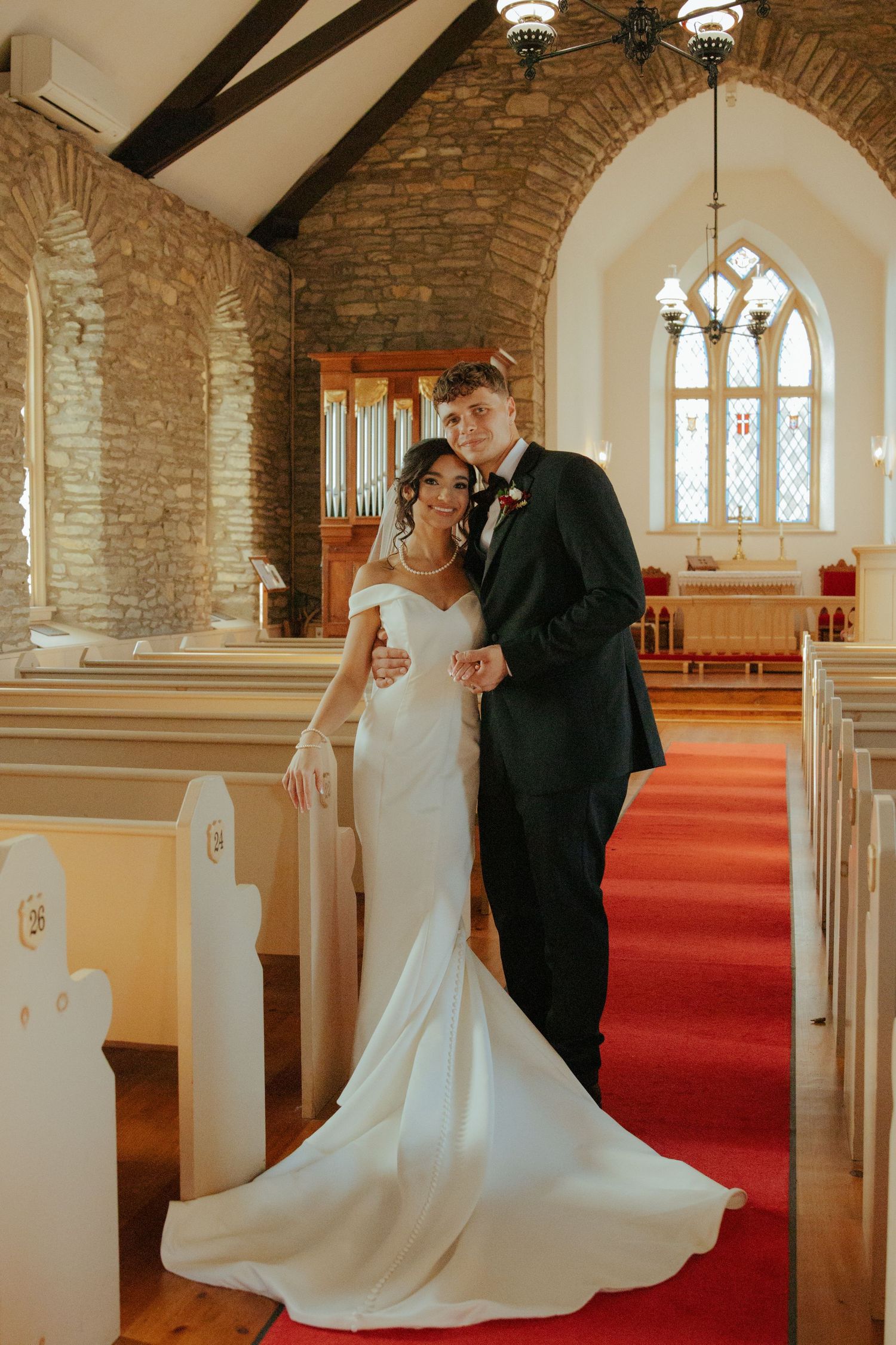 Bride in off-shoulder white gown and groom in black tuxedo pose inside rustic stone church with red carpet aisle.