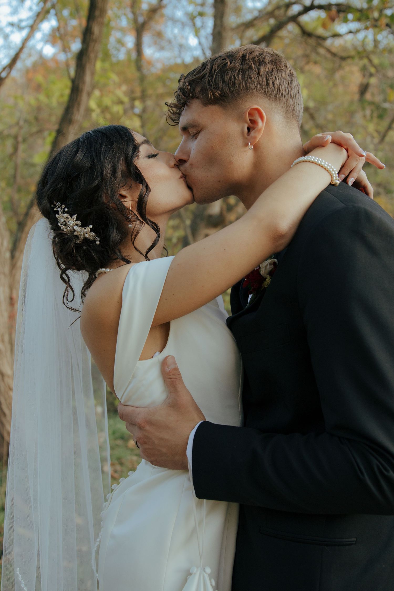 Bride and groom share a romantic kiss outdoors, embracing among autumn trees after their wedding ceremony.