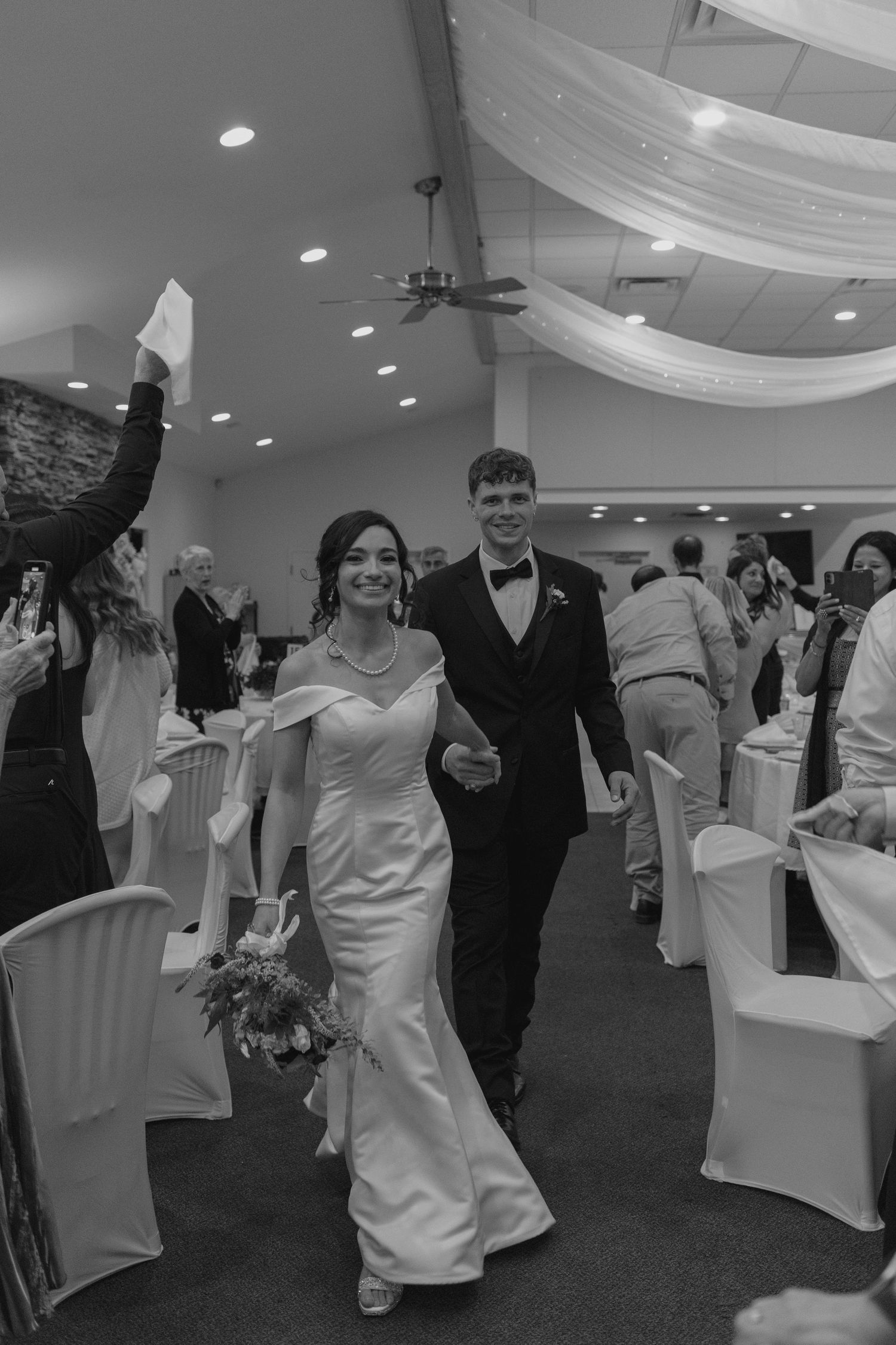 Black and white photo of smiling bride and groom walking through cheering guests at wedding reception.