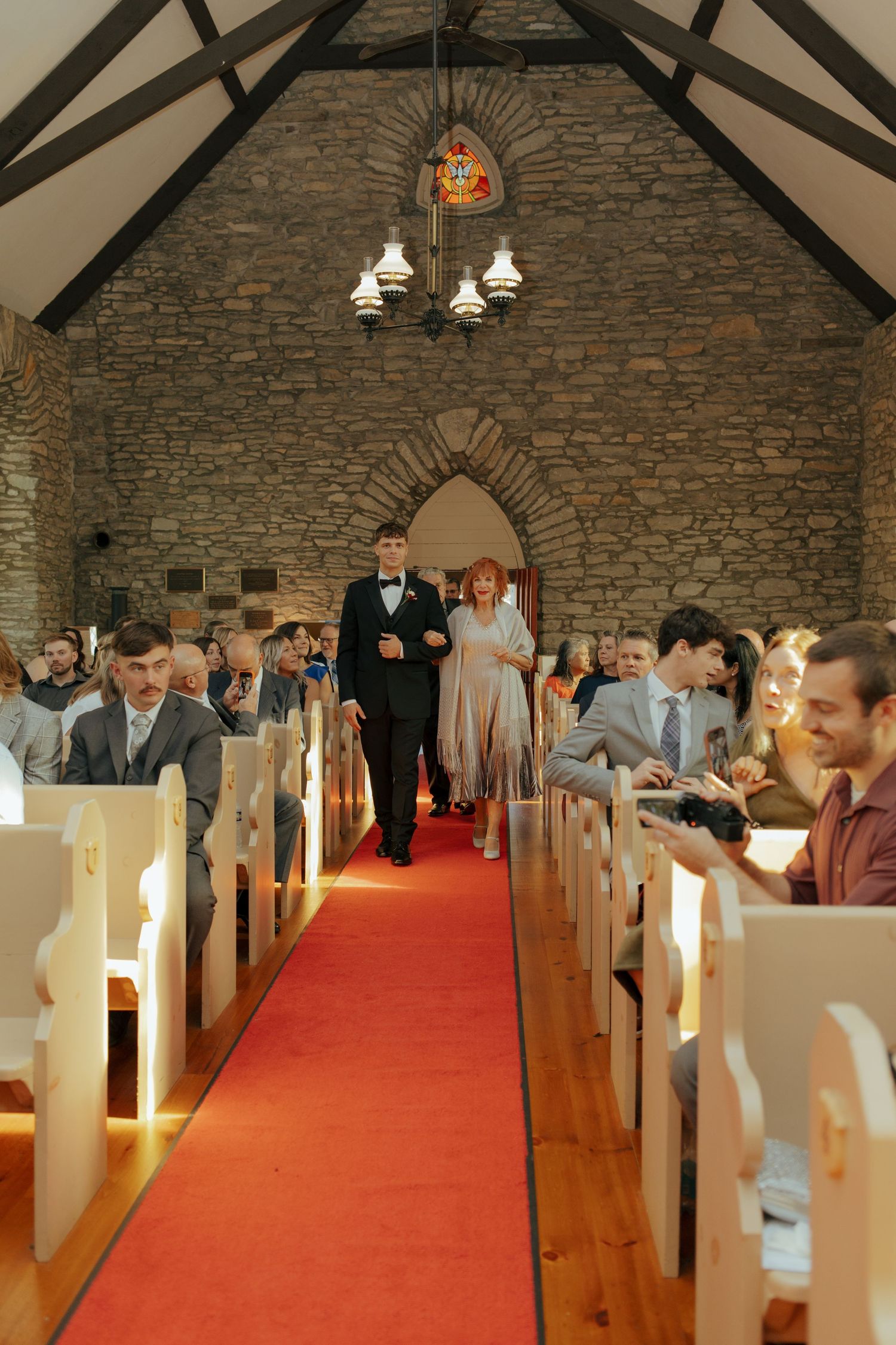 A couple walks down a red carpet aisle in a rustic stone chapel during a wedding ceremony.