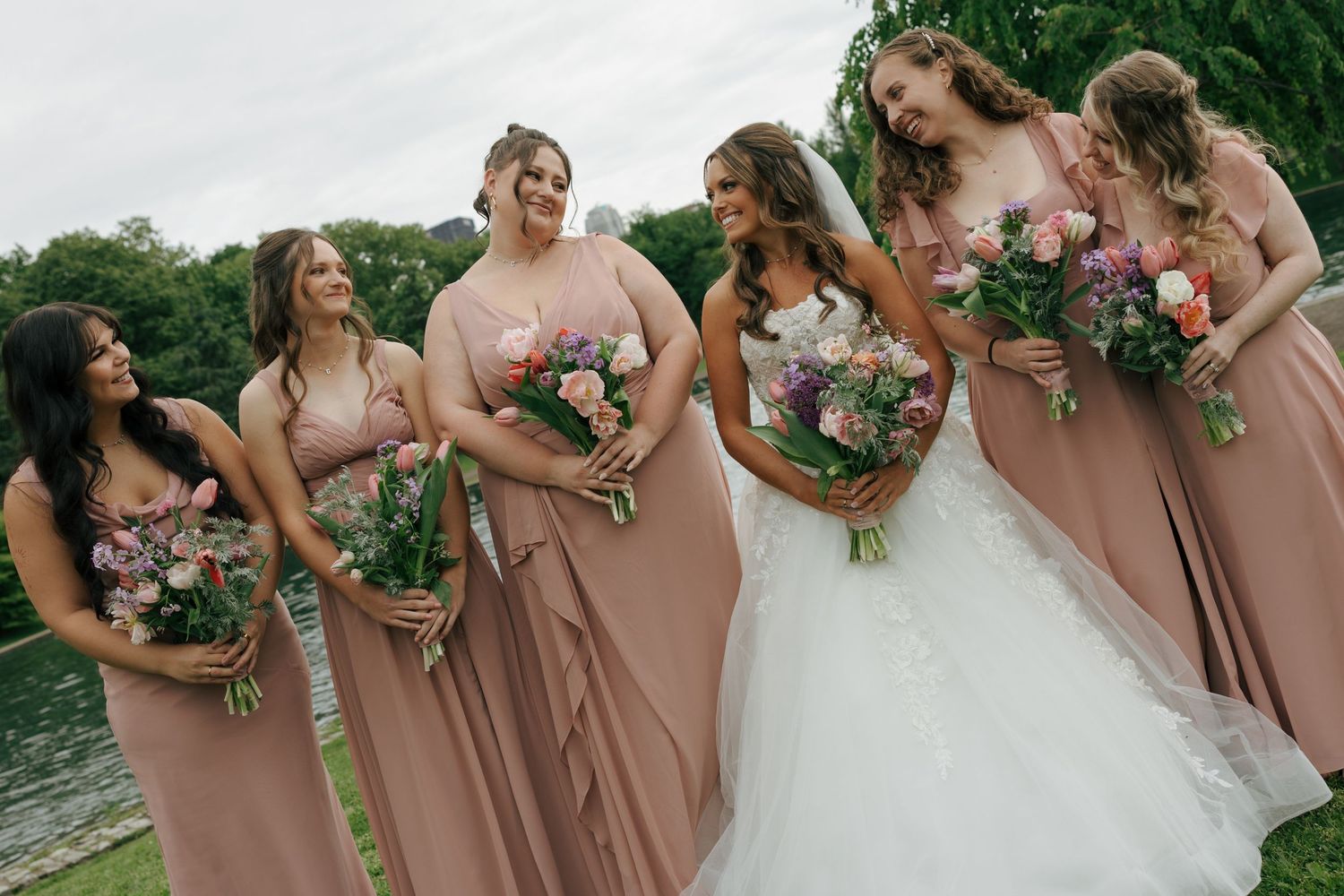 Bride in white gown with bridesmaids in dusty rose dresses holding colorful bouquets outdoors near water.