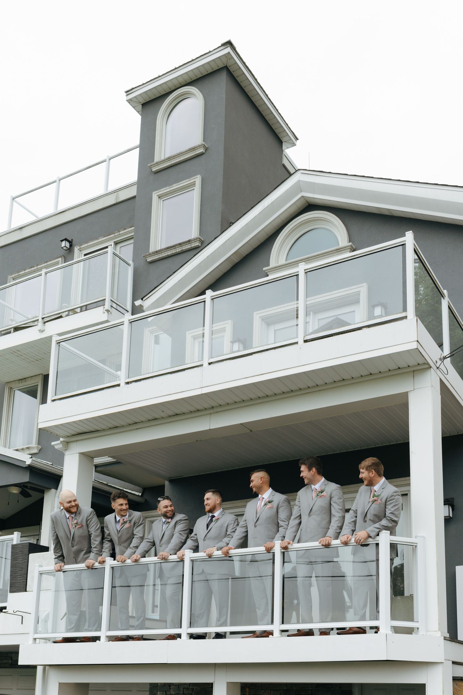 Groomsmen in matching gray suits line up on a white balcony of a gray multi-story venue building.