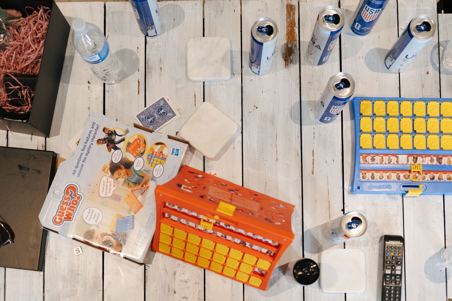 Overhead view of a wooden table with board games, canned drinks, a remote control, and scattered items.