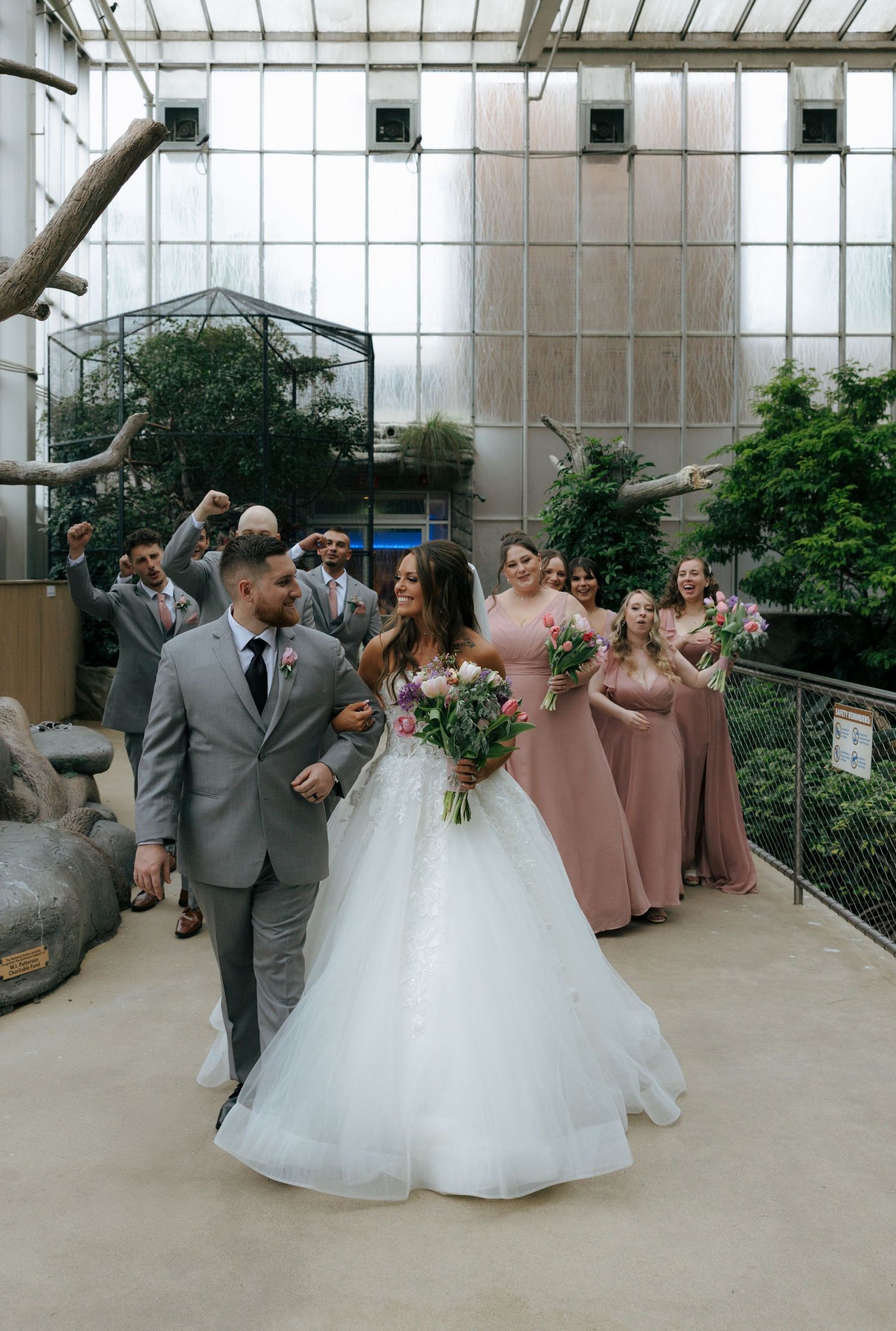 Bride and groom walk together as bridesmaids in pink dresses and groomsmen in gray suits celebrate behind them indoors.