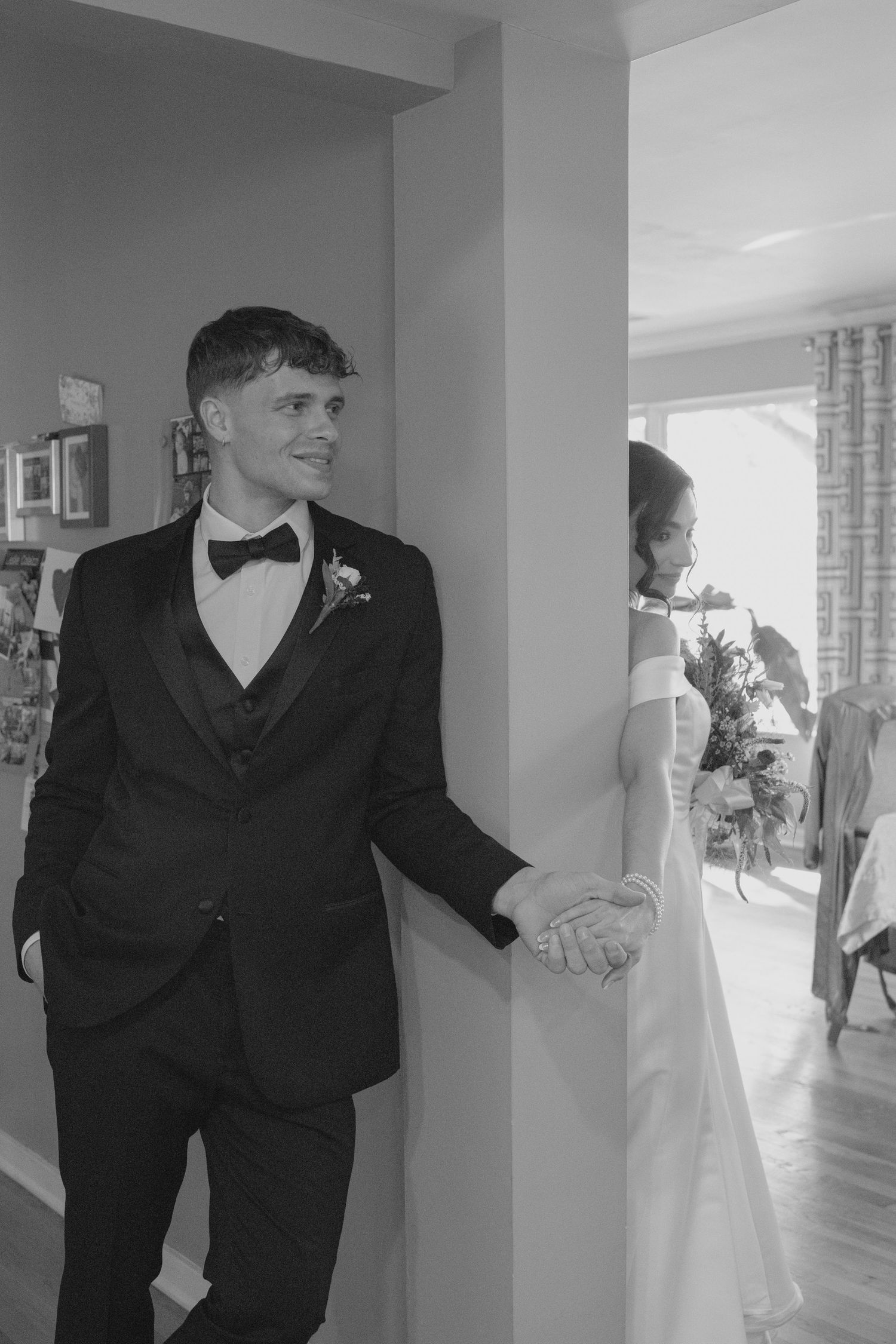 Groom in black tuxedo holds bride's hand around a doorway in a romantic black and white wedding photo.