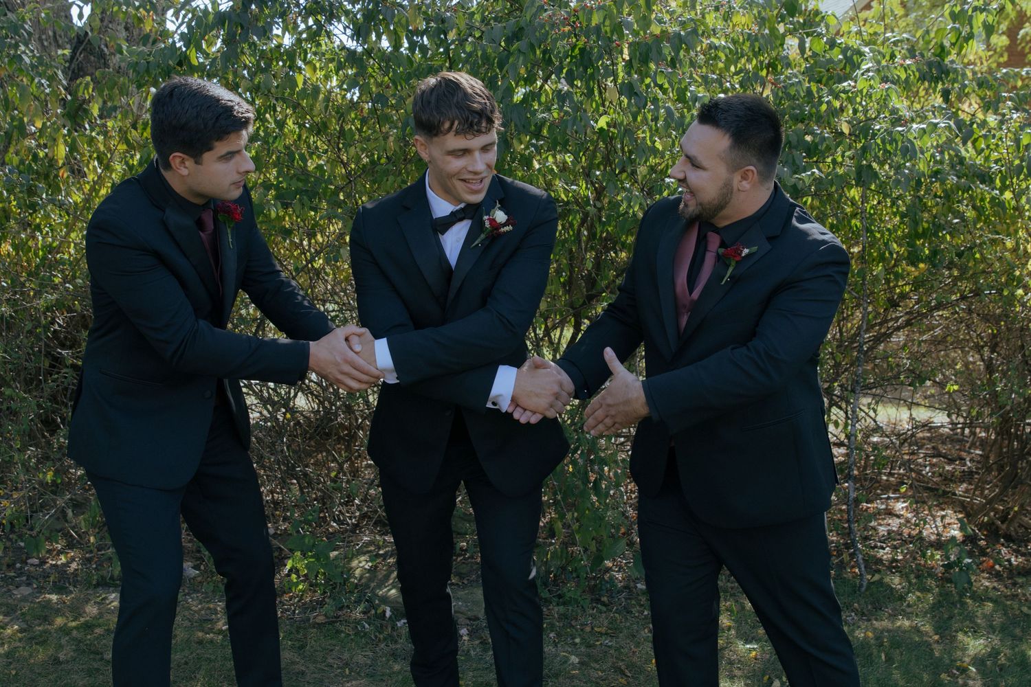 Three young men in black suits shake hands outdoors among green trees at a formal event.