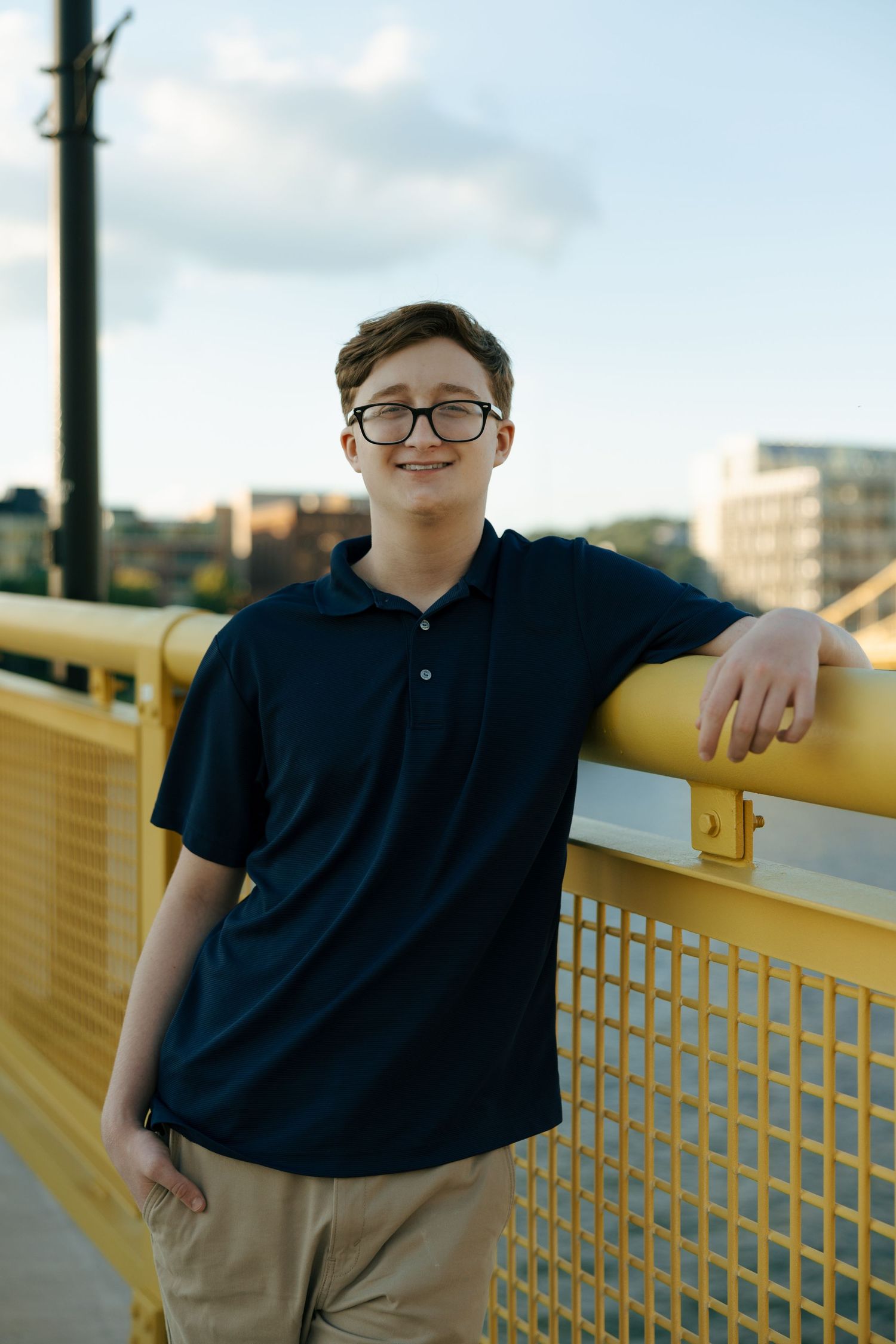 Teen boy in navy polo and glasses smiling on a yellow bridge railing in an urban setting.