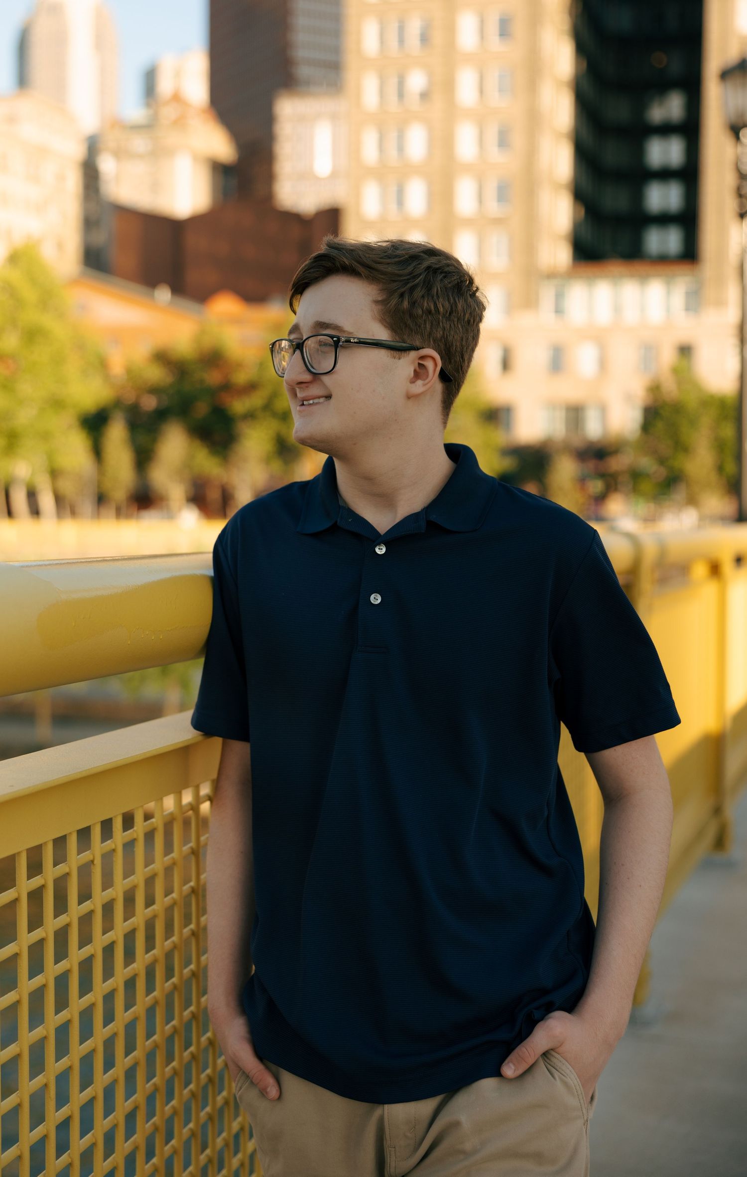 Teen boy in navy polo shirt and glasses smiling near yellow bridge railing with Pittsburgh skyline.
