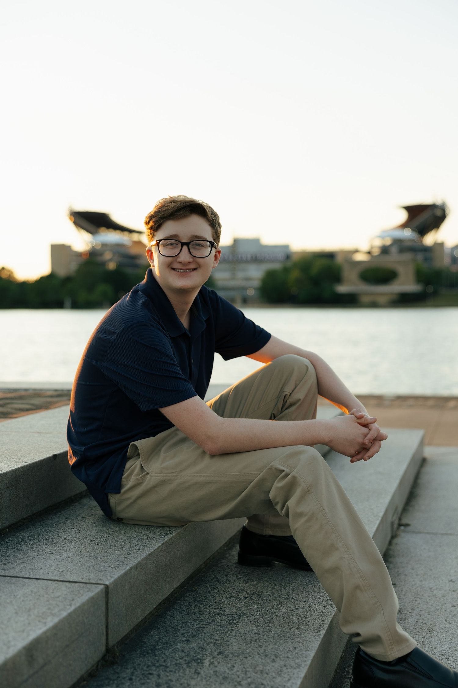 Smiling young man in navy polo sitting on stone steps by river with stadium in background at sunset.