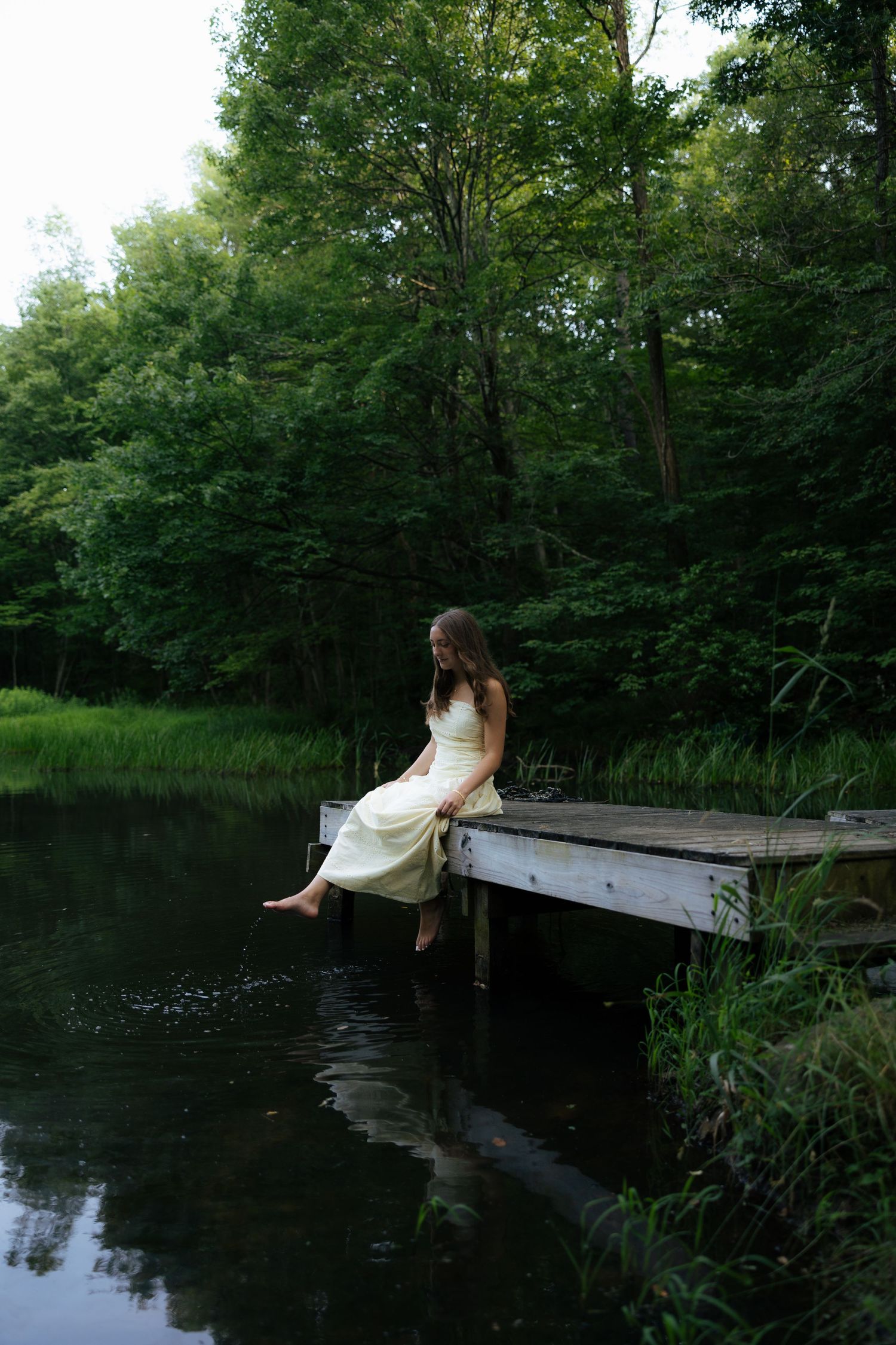 Young woman in white dress sitting on a wooden dock with feet dangling over a tranquil forest pond.