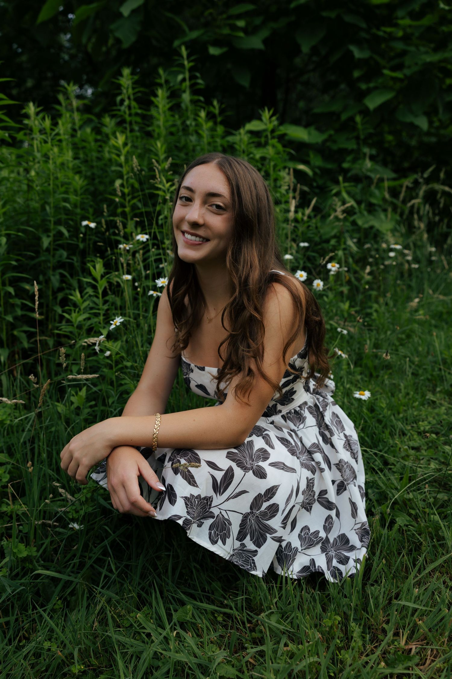 Young woman in floral sundress smiling while crouching in lush green meadow with wildflowers.