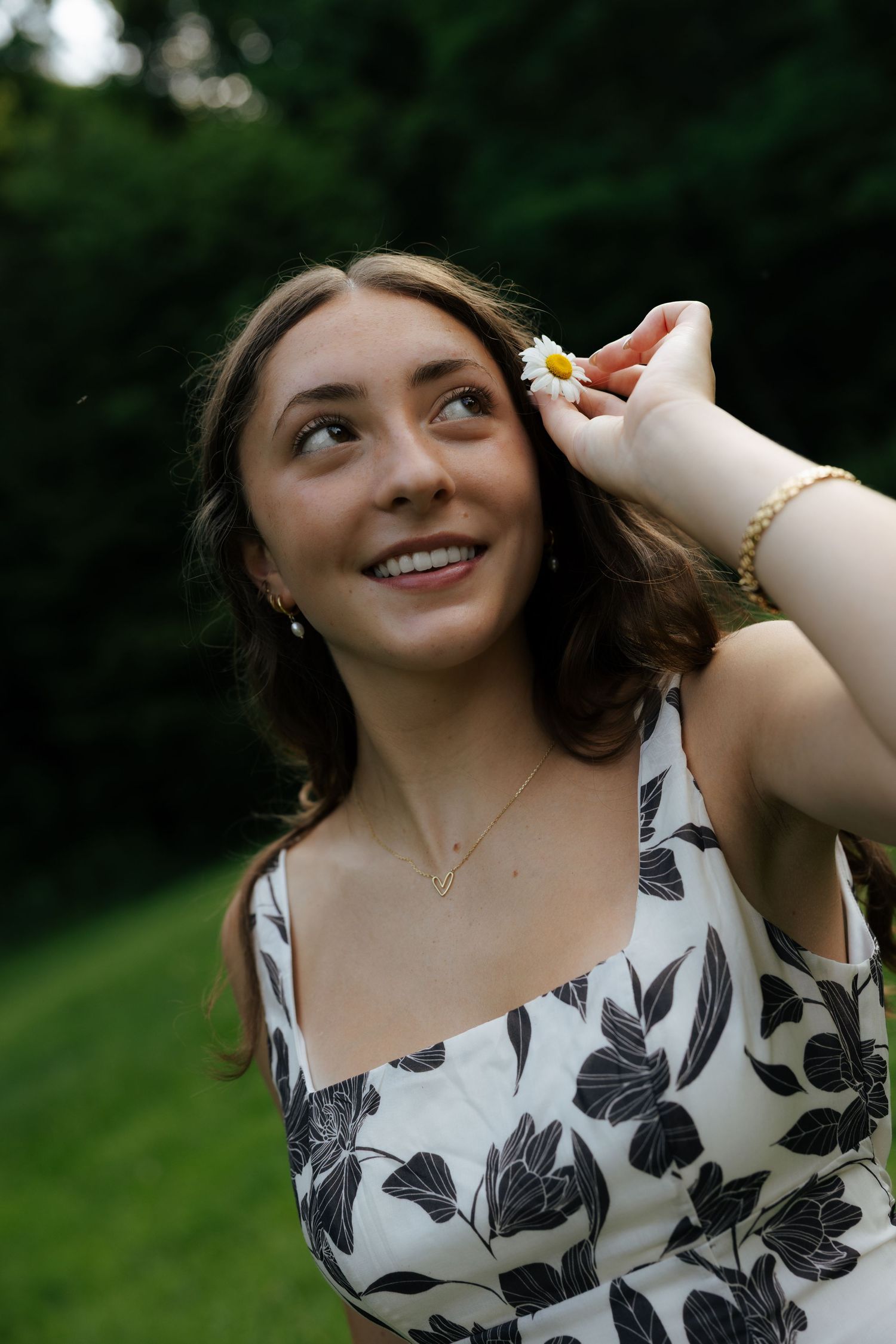 Young woman smiling outdoors placing a daisy flower in her hair wearing a floral dress and gold jewelry.