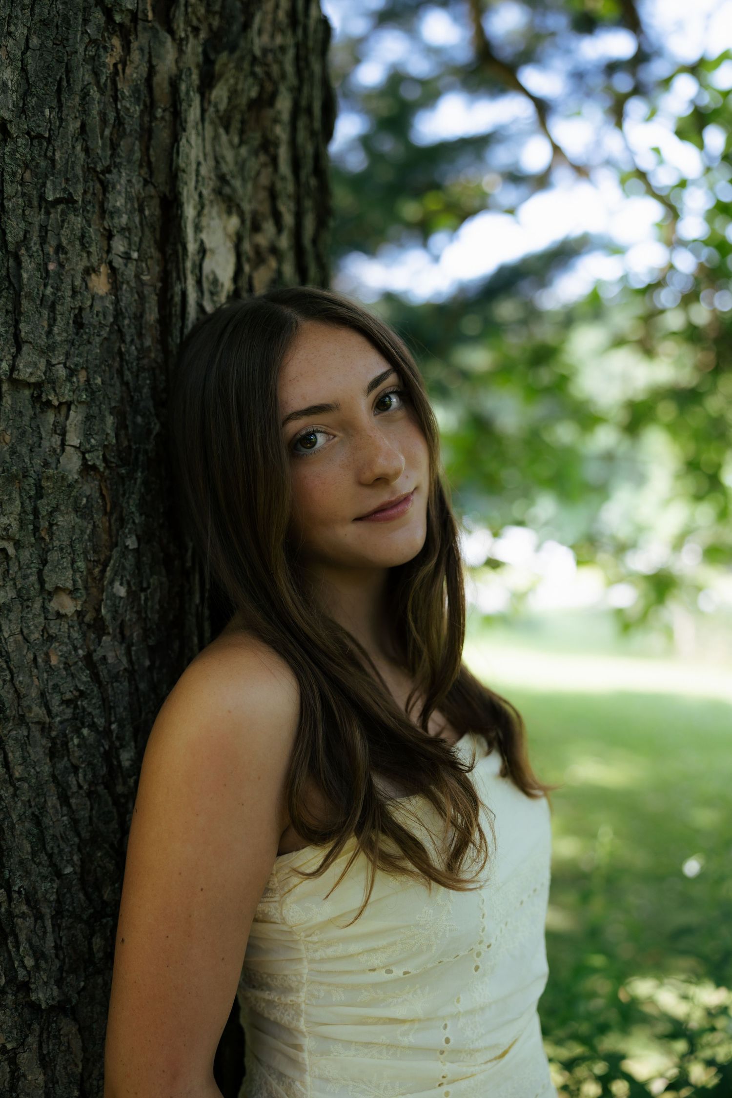 Portrait of brunette teen girl leaning against tree trunk wearing white strapless dress outdoors.