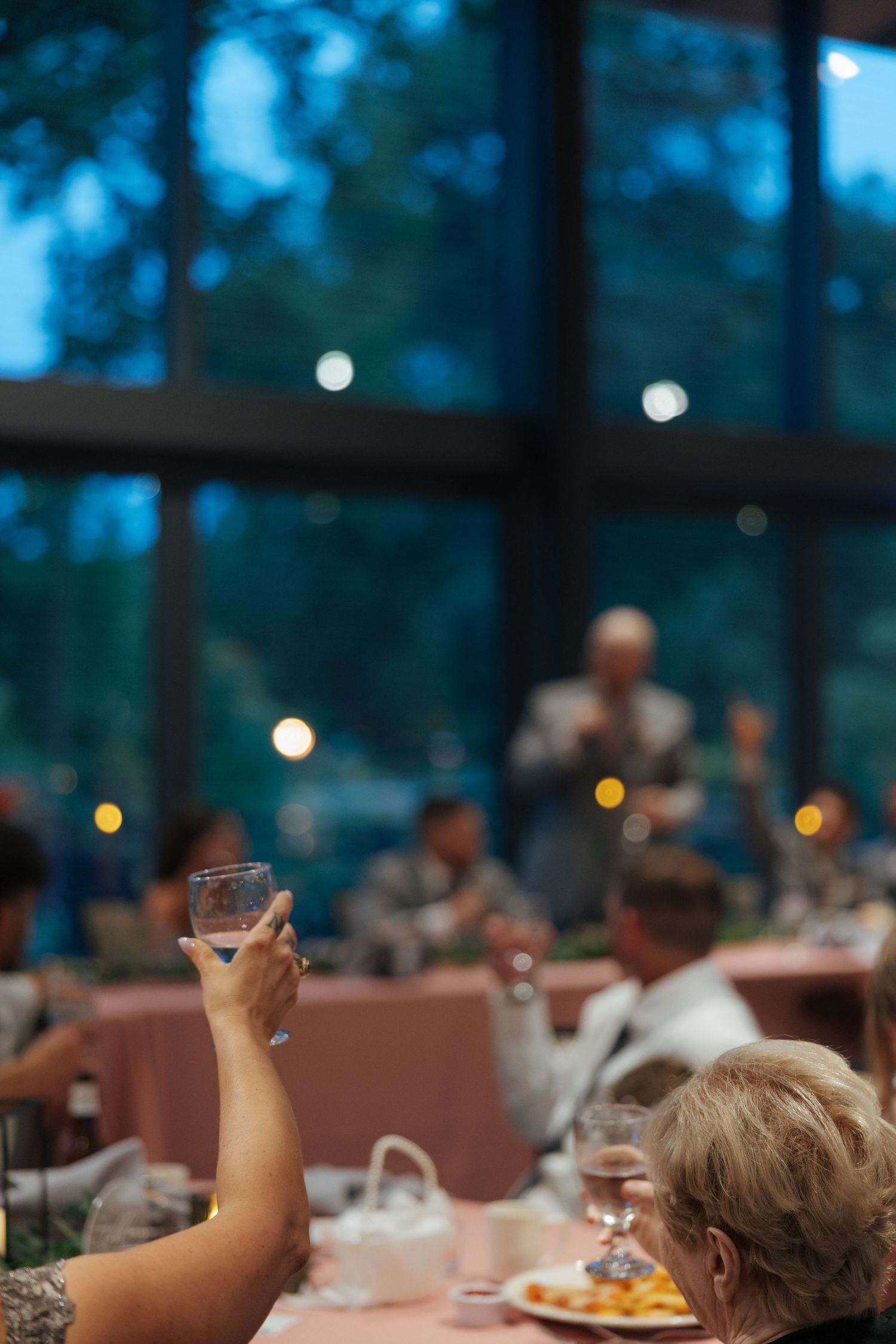 Wedding guest raises glass for champagne toast during evening reception with bokeh lights in background.