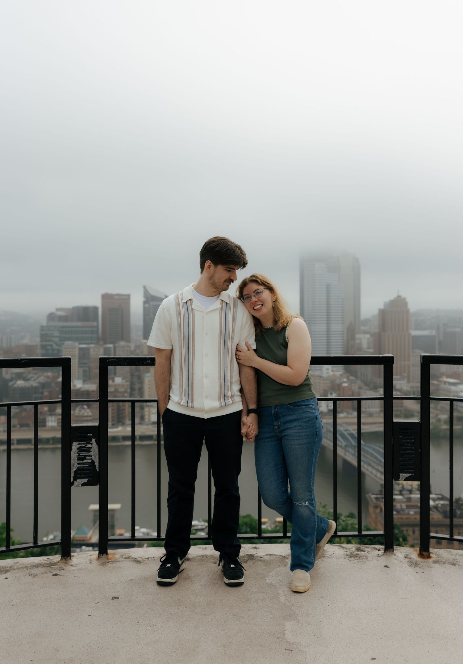 A happy couple stands on a rooftop overlook with the Pittsburgh skyline behind them on a cloudy day.