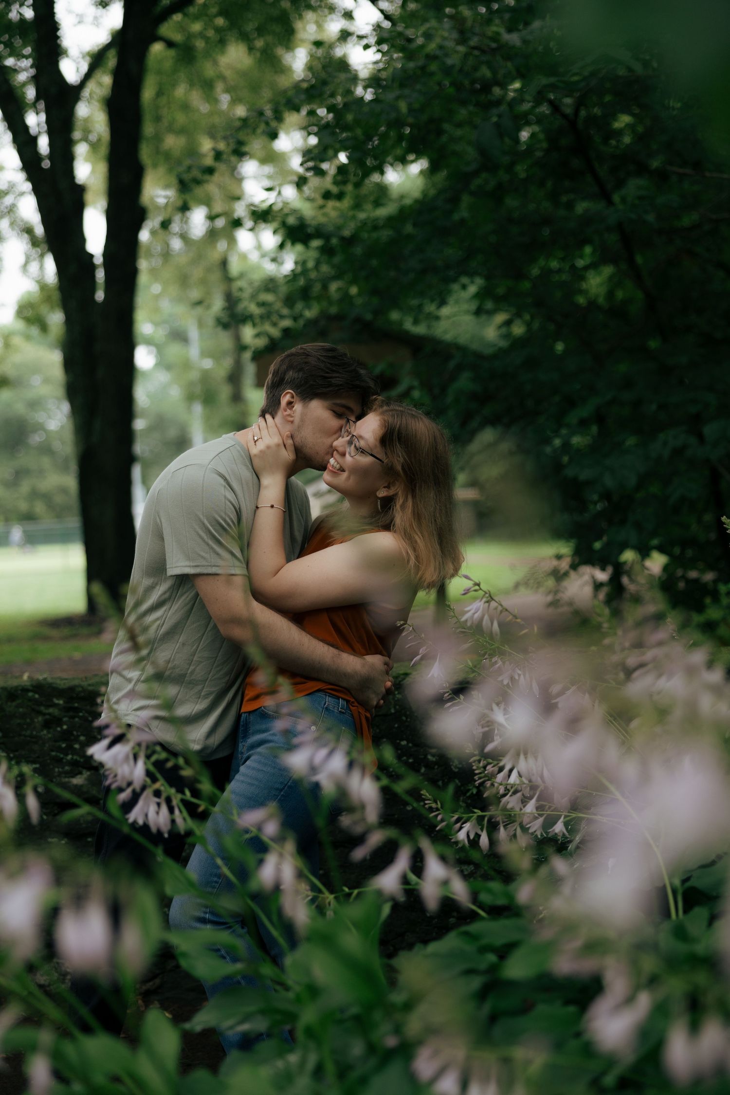 Couple embracing and kissing among lush green foliage and white flowers in a serene park setting.