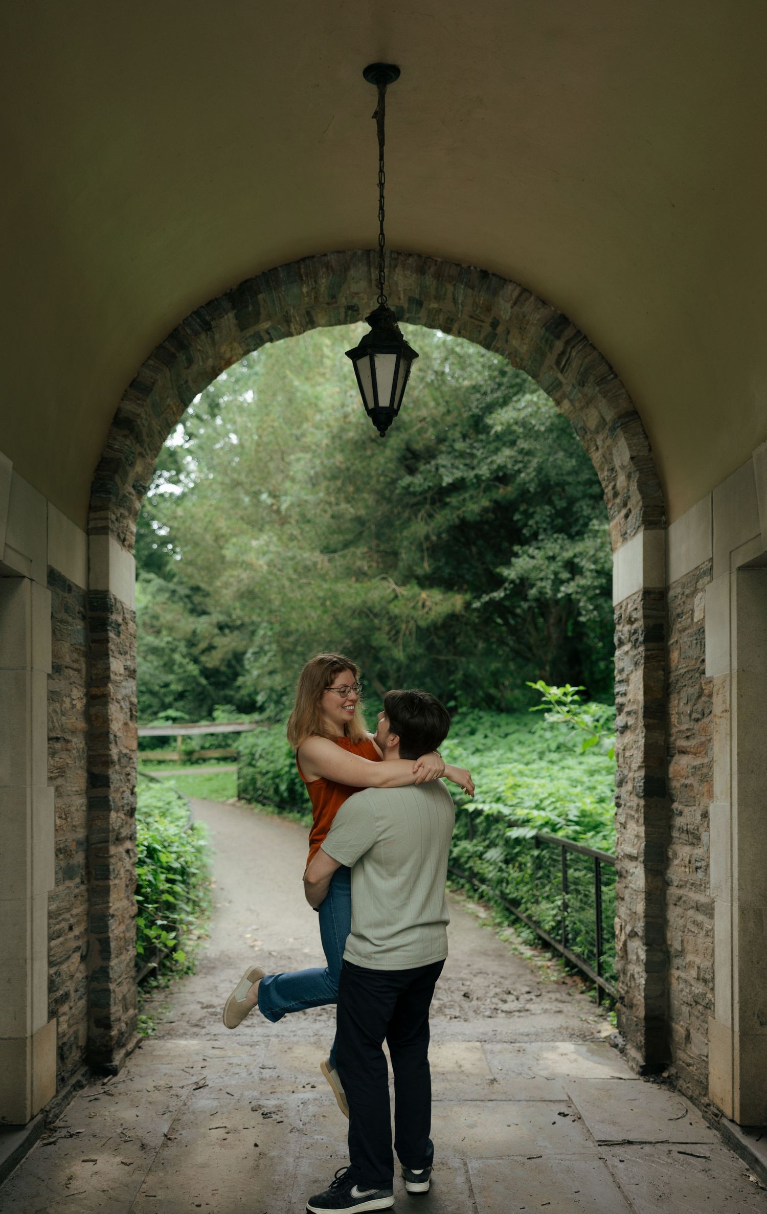 Couple sharing a joyful embrace under a stone archway with lush green garden path in the background.