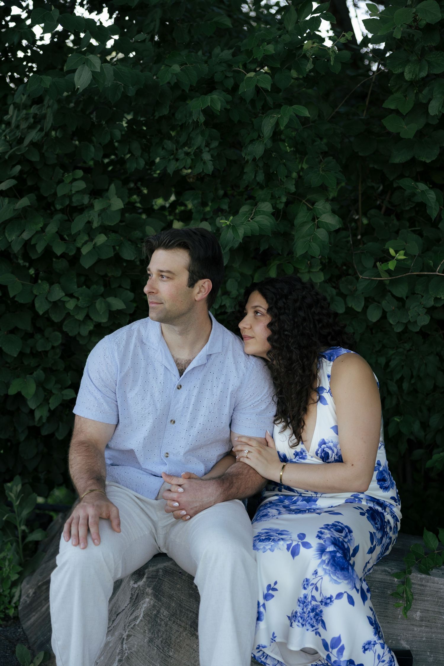 A couple sits on a stone bench surrounded by lush green foliage, gazing into the distance.