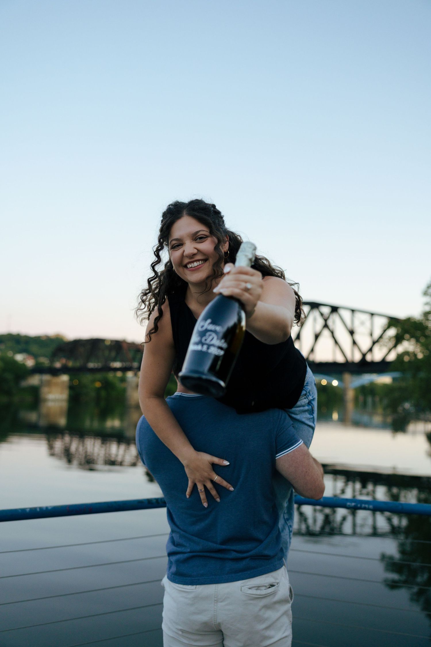 Joyful woman being carried piggyback by man holding champagne bottle near waterfront bridge at sunset.
