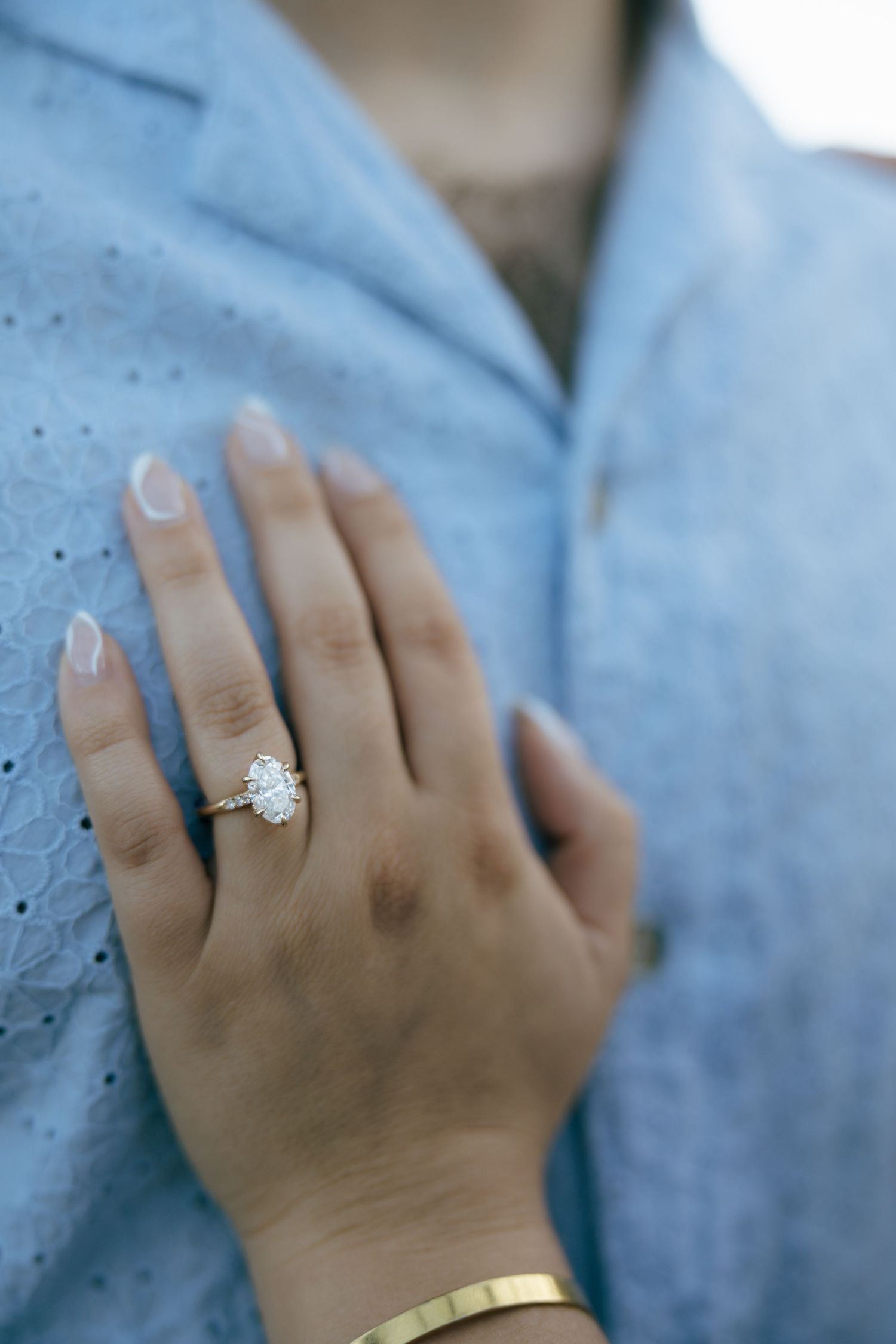 Close-up of an oval diamond engagement ring on a woman's hand resting on a man's light blue shirt.