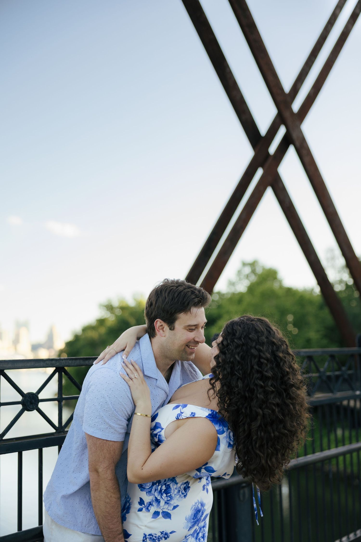 A smiling couple leans in for a kiss on an old iron bridge under a clear blue sky.