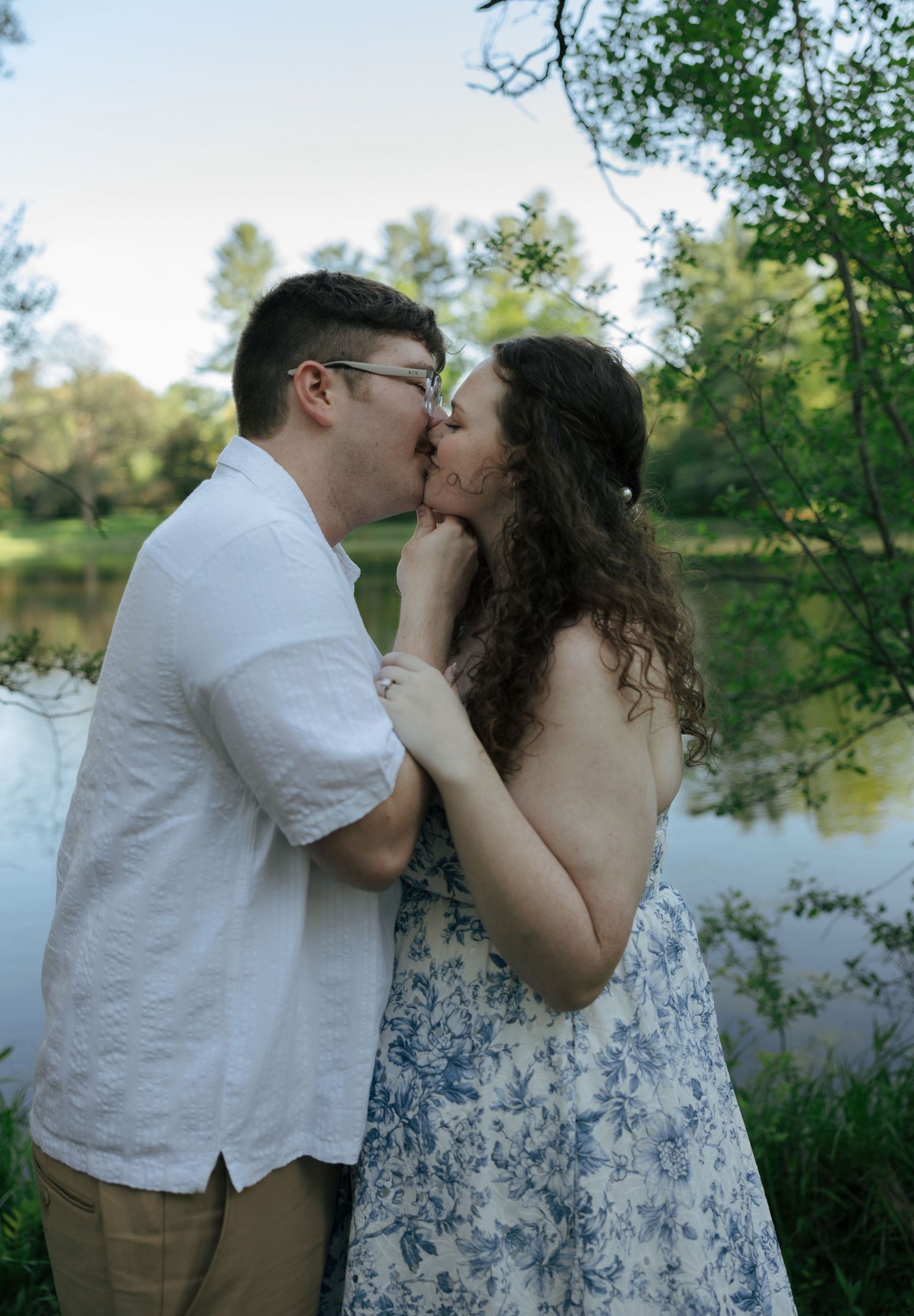Couple sharing a romantic kiss by a serene pond surrounded by lush green trees outdoors.