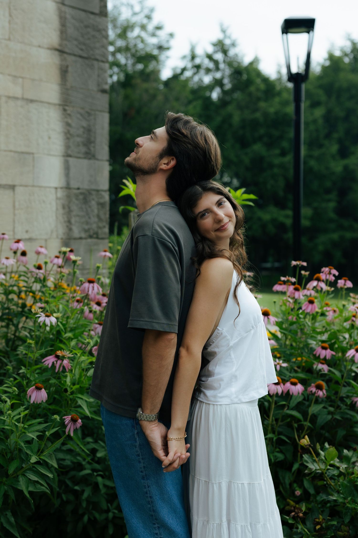 Couple holding hands in a garden, woman in white dress leaning on mans back near pink flowers.