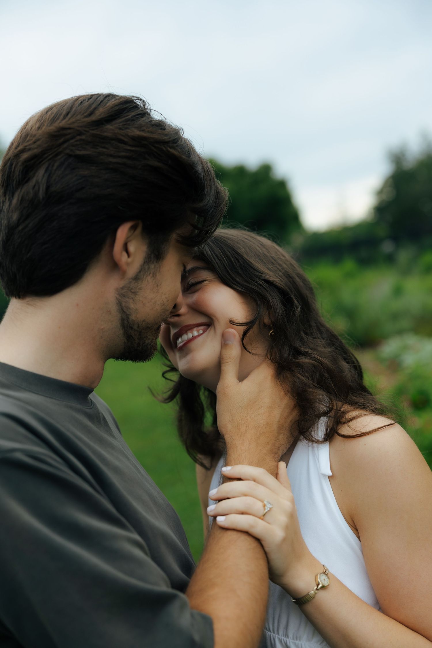 A happy couple shares an intimate moment outdoors, man gently cupping womans face as she laughs joyfully.