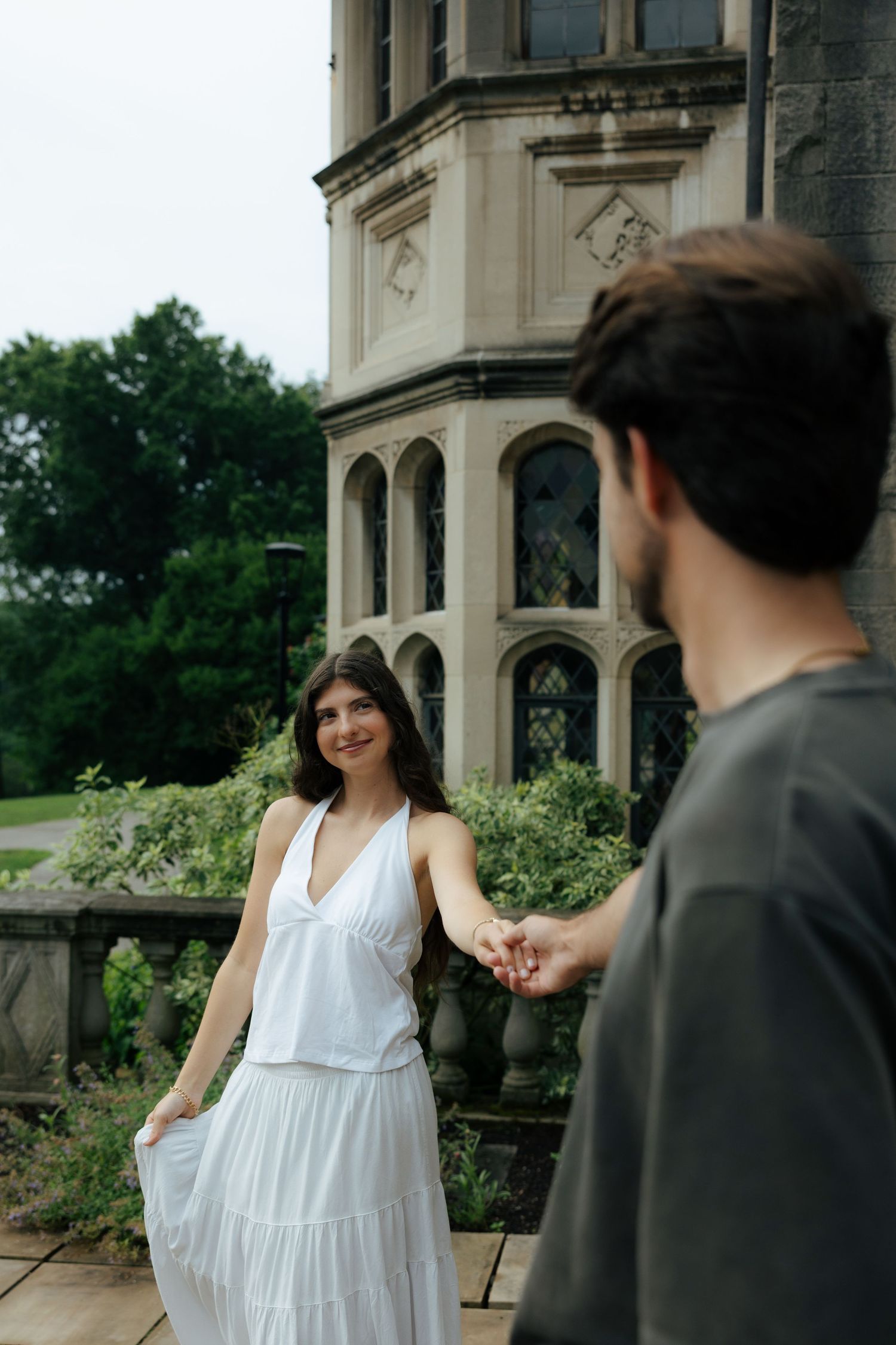 Woman in white dress holding man's hand, smiling in front of a grand stone building.