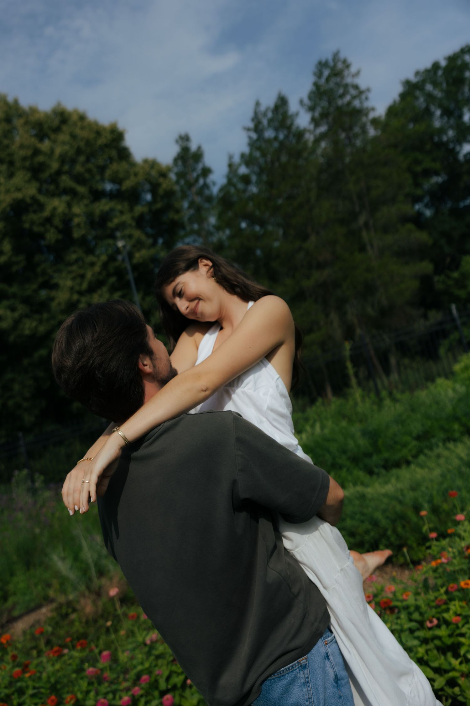 Man lifting woman in white dress in a garden, sharing a romantic moment among flowers and trees.
