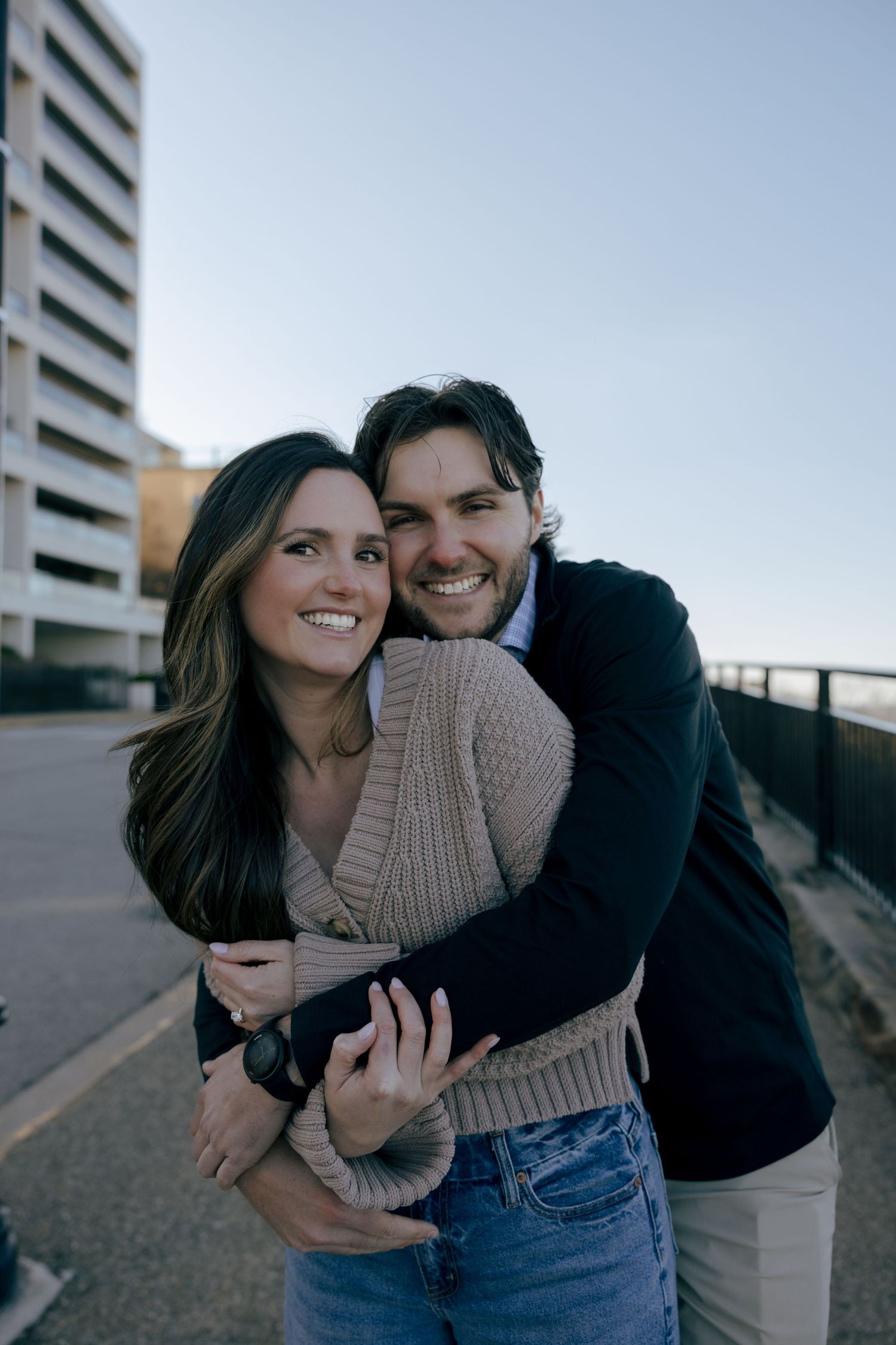 A smiling couple embracing outdoors on a urban walkway, woman in beige sweater and jeans, man in dark blazer.