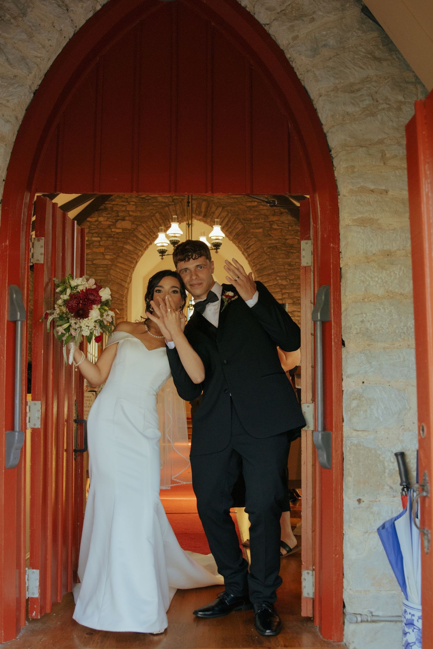 Wedding couple poses playfully in front of rustic red archways and stone walls.