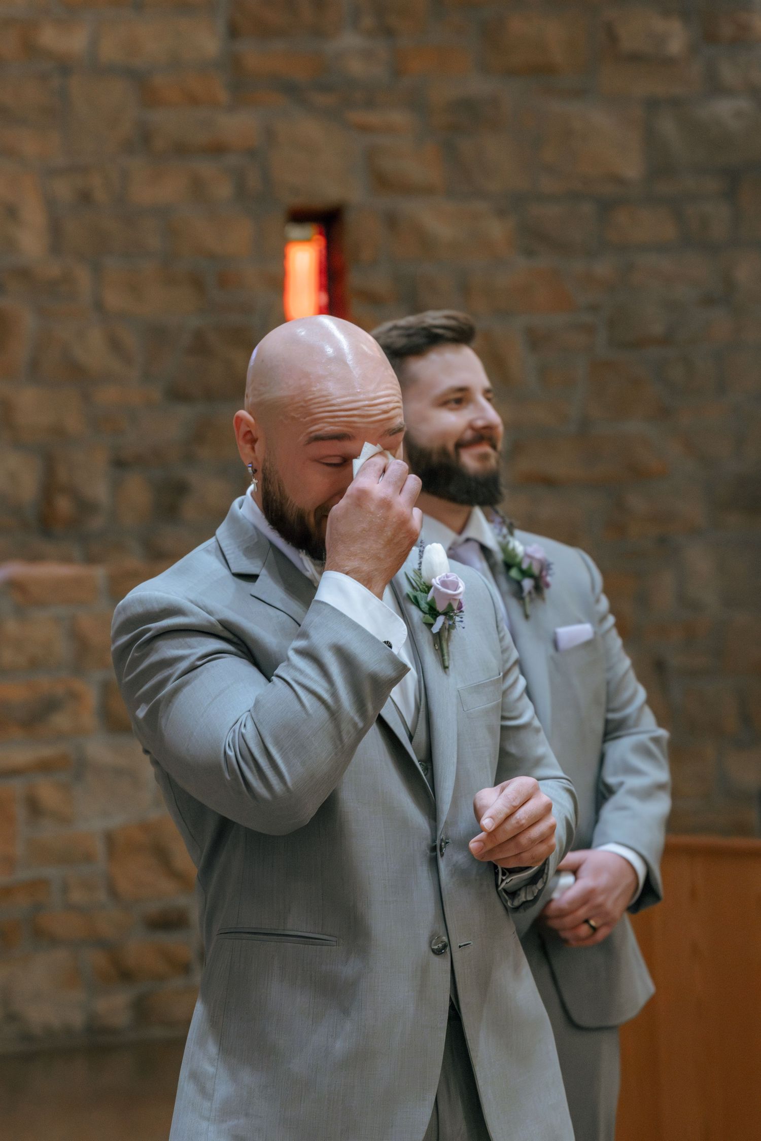 Two groomsmen in light gray suits share an emotional moment during a wedding ceremony against a brick wall.