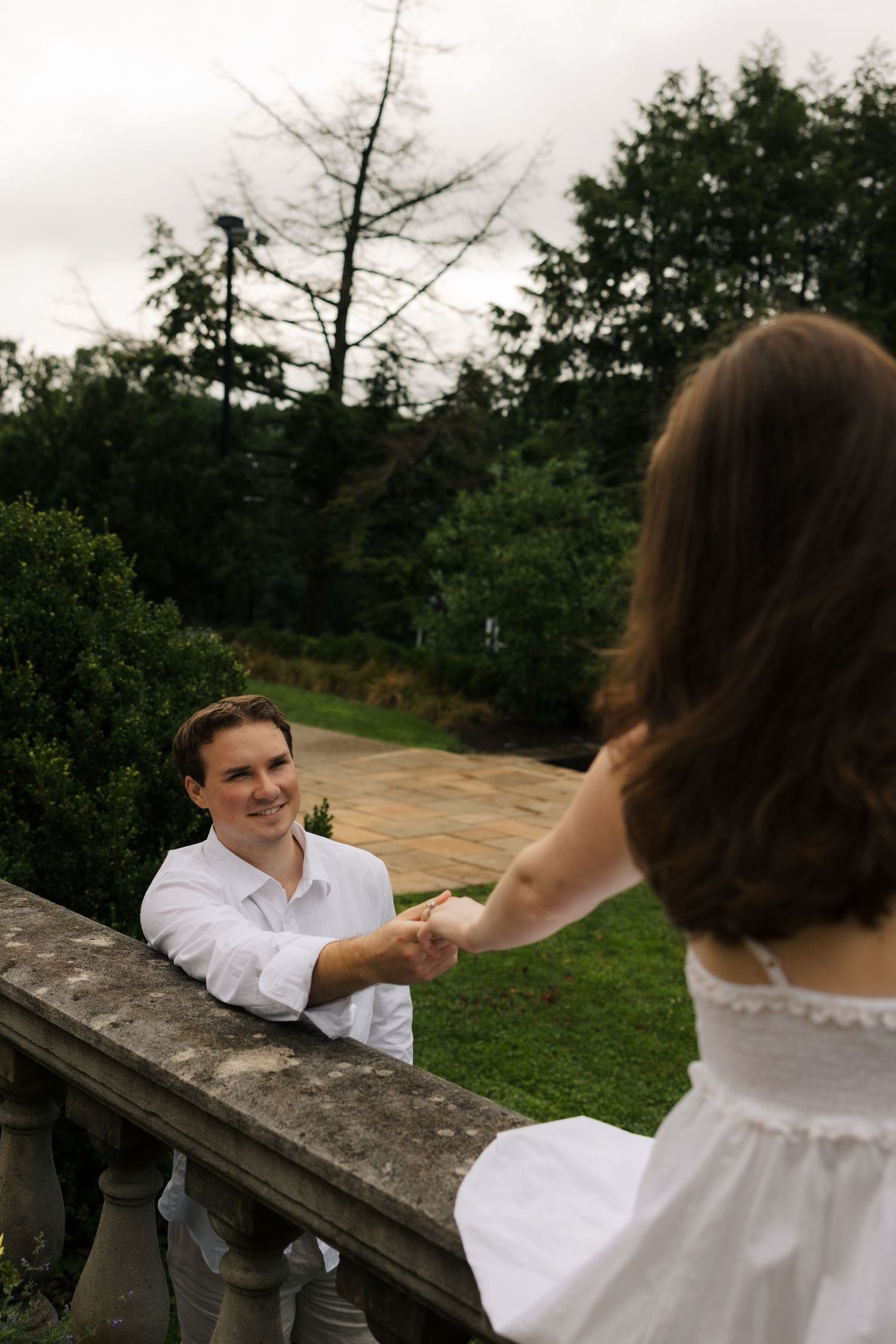 A smiling man in a white shirt holds hands with a woman in a white dress over a stone balustrade in a lush garden.