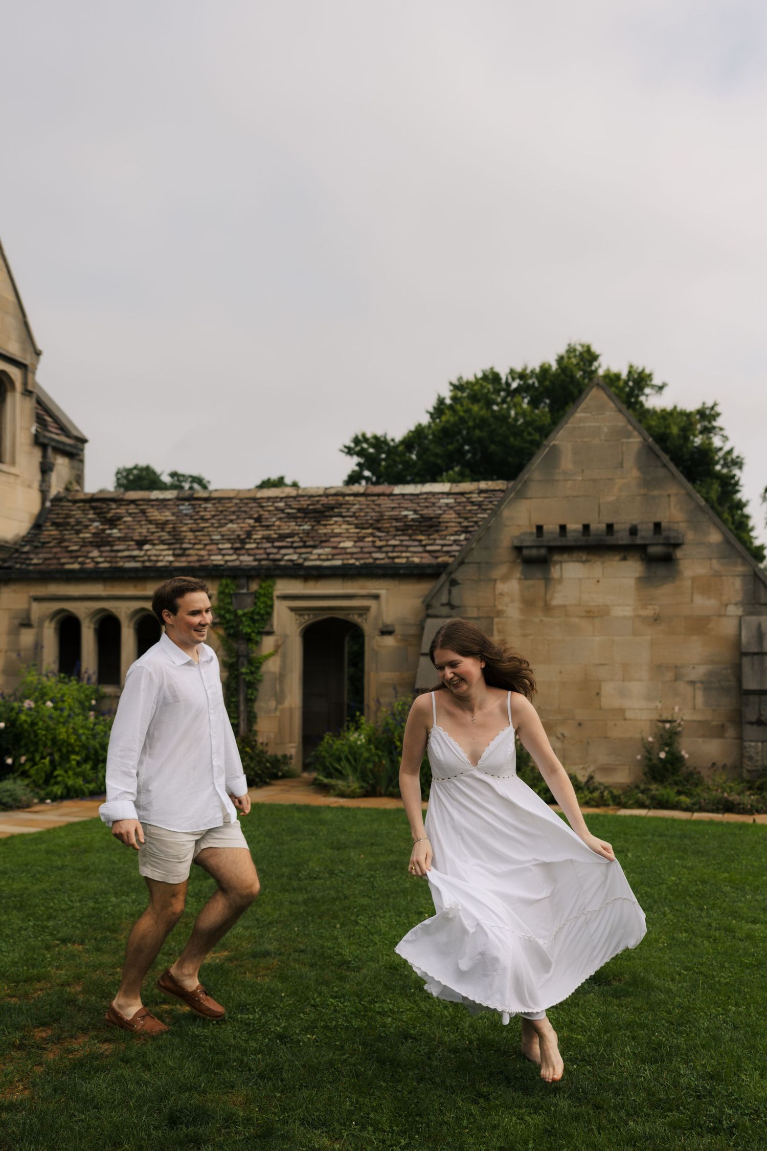 Couple in white outfits joyfully running together on a green lawn in front of a historic stone manor.