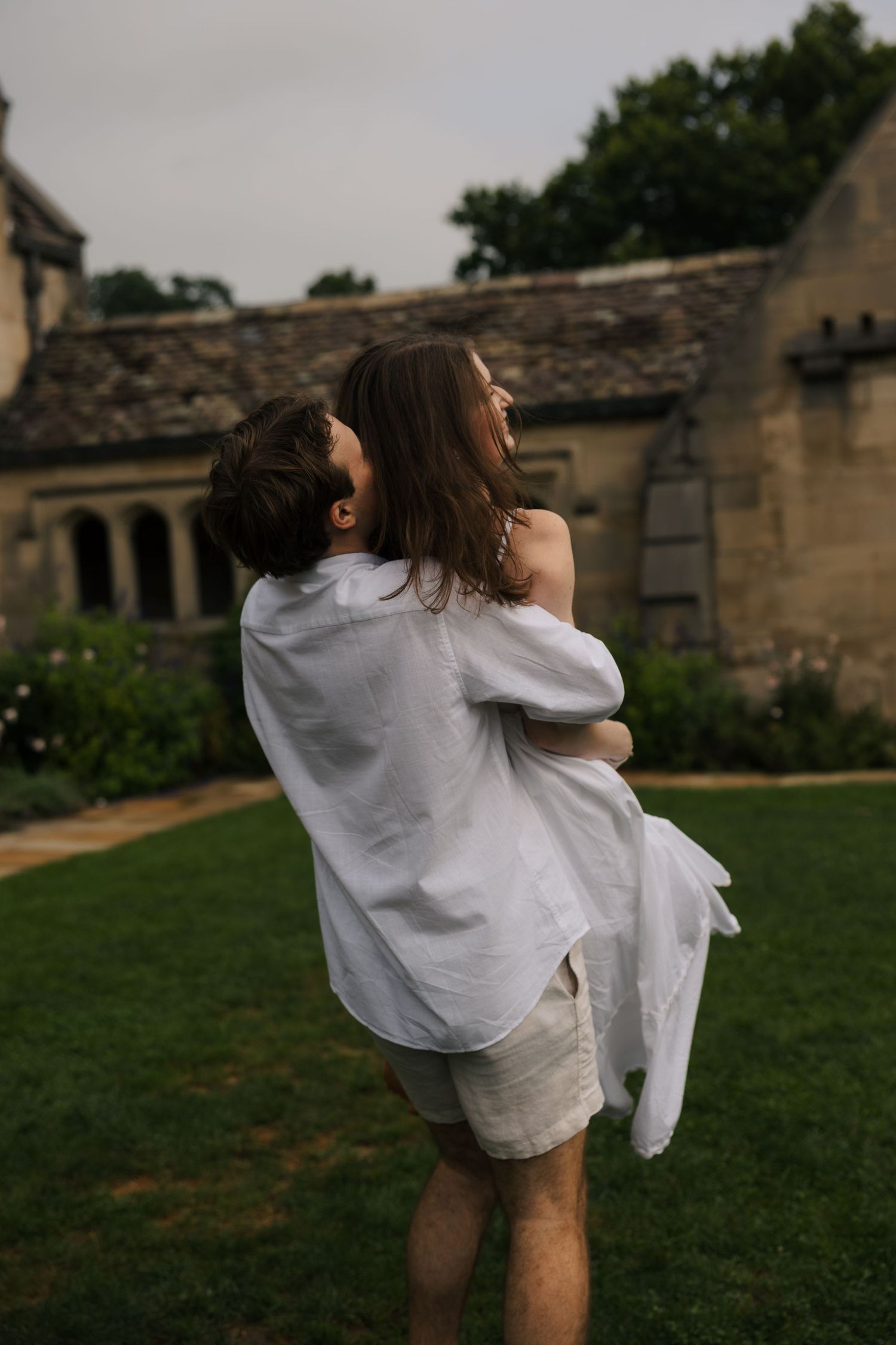 Man in white linen shirt lifts laughing woman in his arms outside historic stone building.