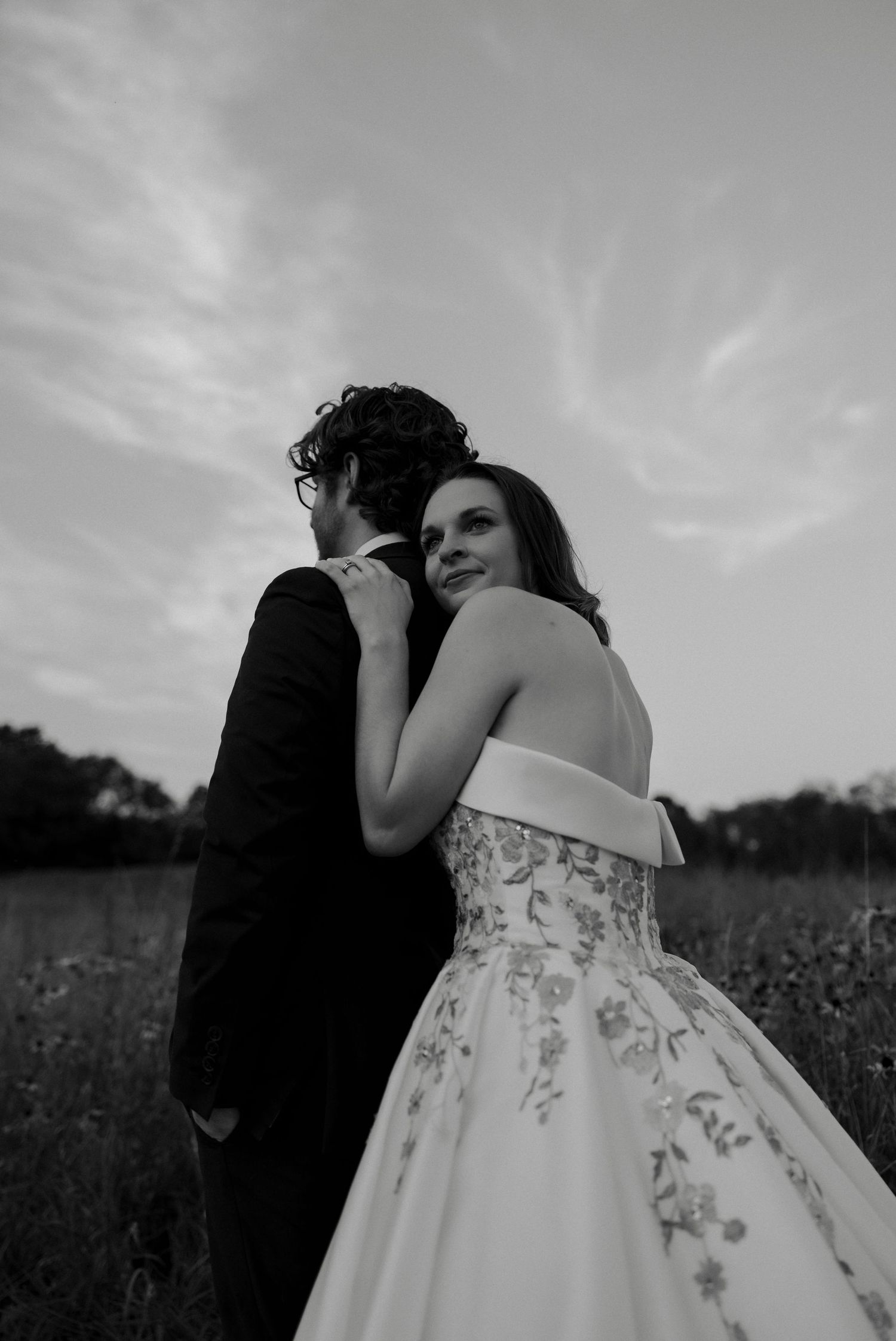 Black and white wedding portrait of bride in strapless floral gown embracing groom in open field at sunset.