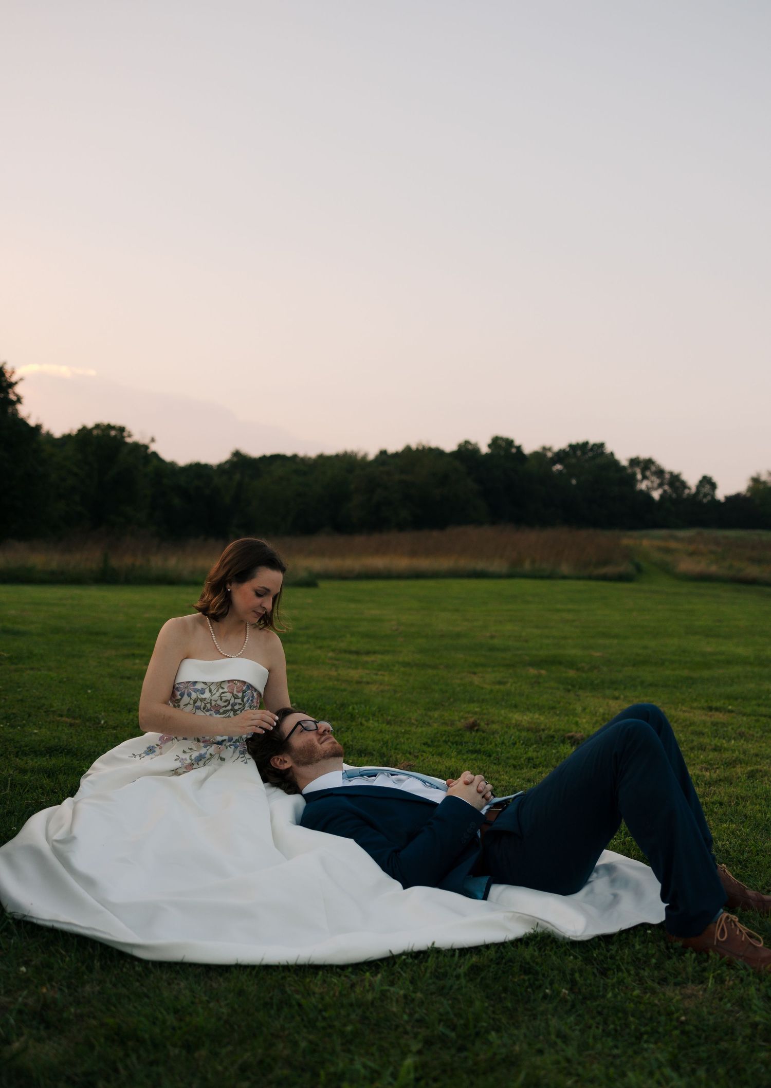 Bride in strapless floral gown sits on grass while groom lays in her lap during outdoor sunset wedding.