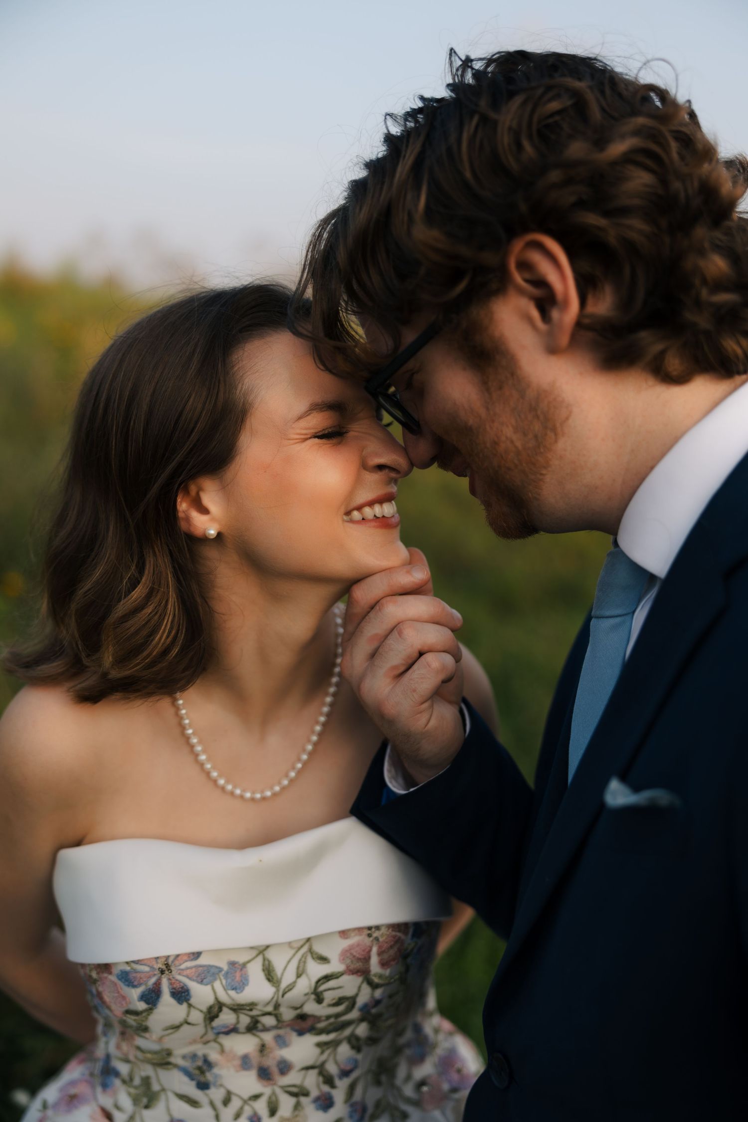 Bride and groom touching foreheads romantically, smiling, bride wearing floral wedding gown and pearl necklace outdoors.