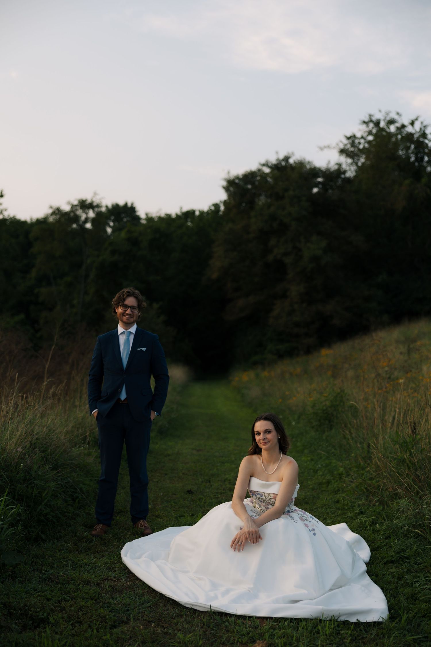 Bride in floral ballgown sitting on grass path while groom in navy suit stands behind her at dusk.
