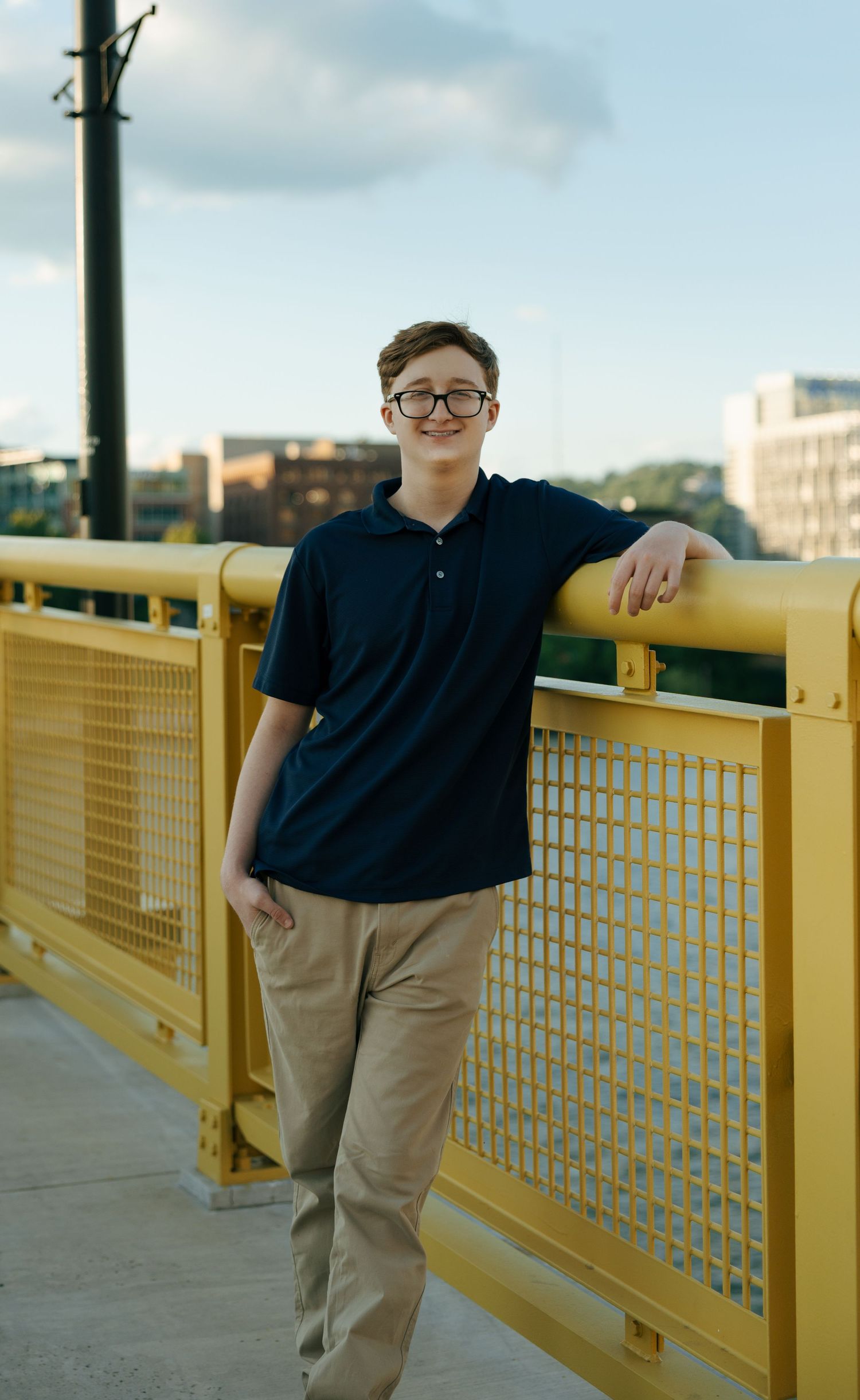 Teen boy in navy polo and khakis leaning on yellow bridge railing with city skyline background.