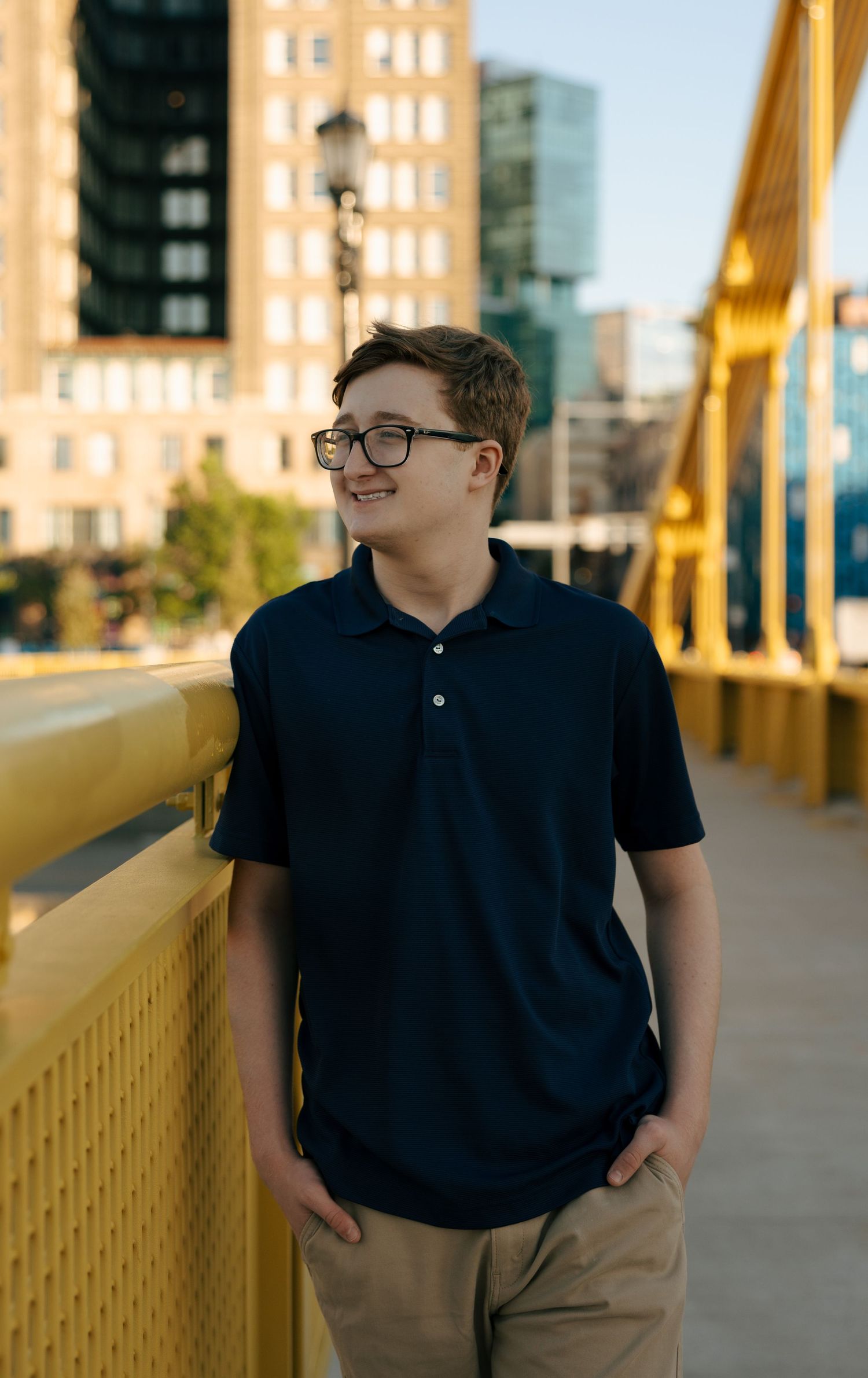 Smiling teenage boy in navy polo shirt and glasses standing on a yellow bridge with city buildings in background.