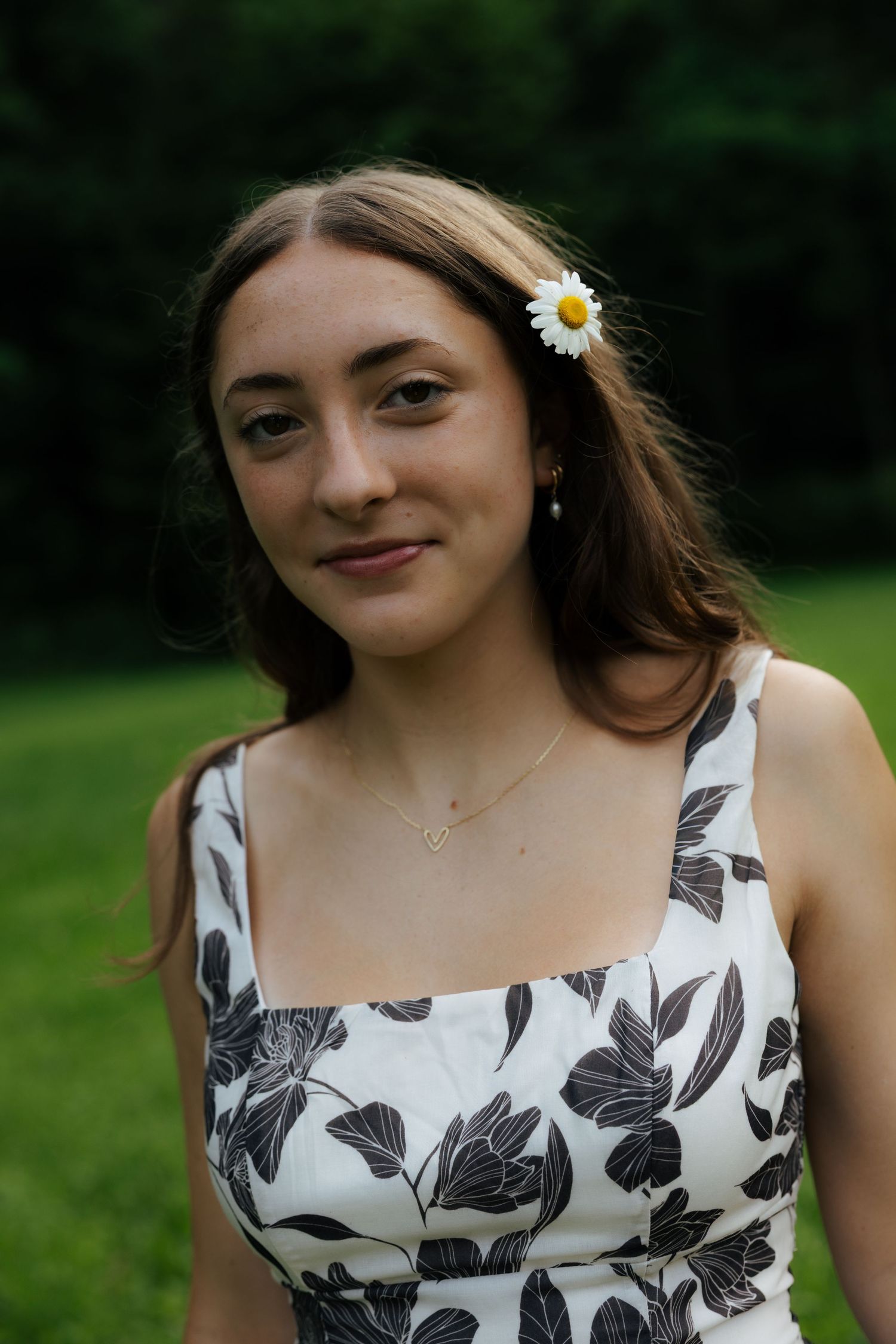 Smiling young woman with daisy flower in hair wearing floral dress outdoors in green park setting.
