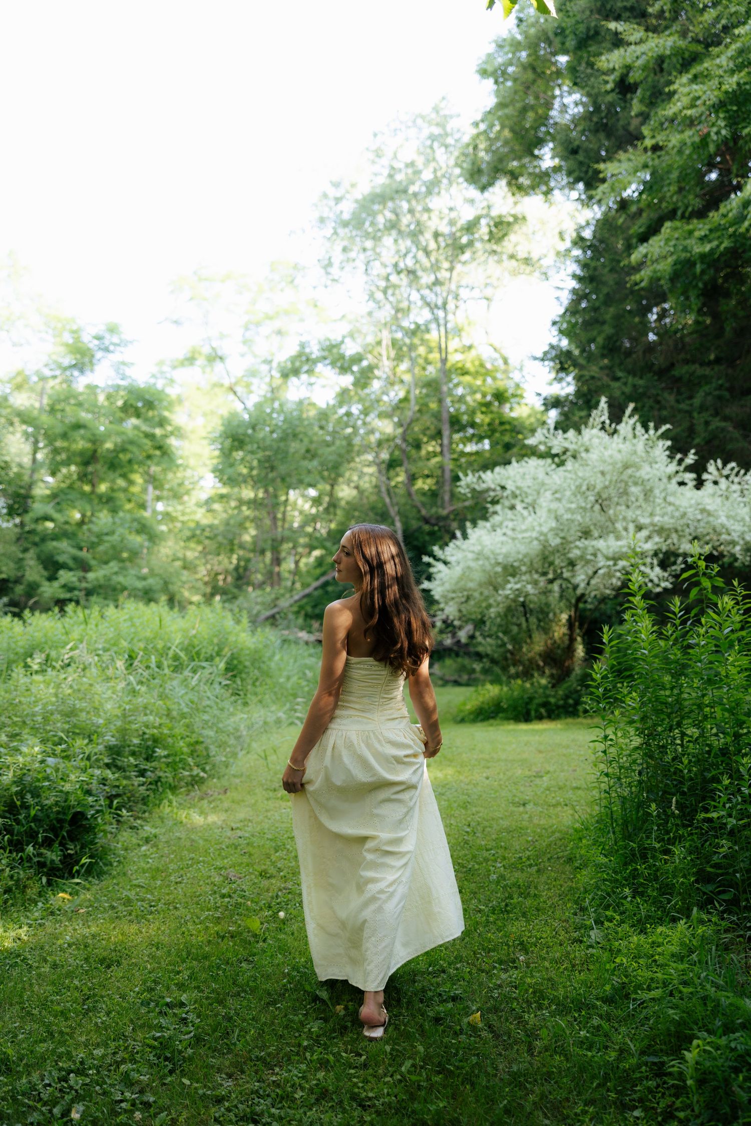 Young woman in elegant cream maxi dress walking through lush green garden path surrounded by blooming shrubs.