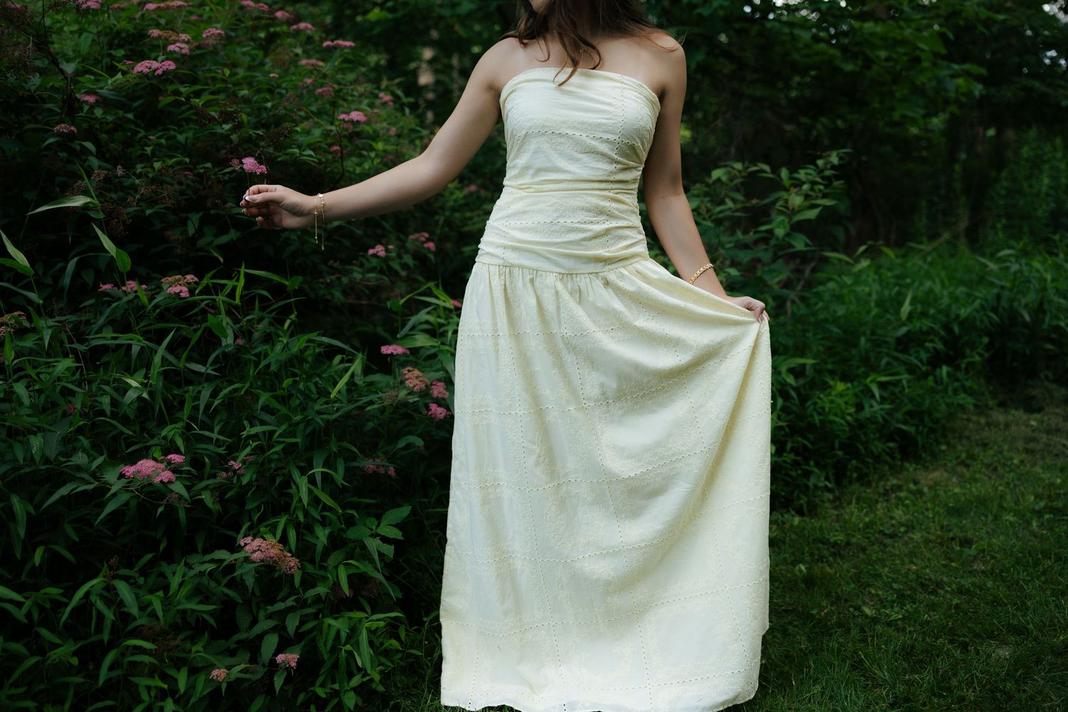 Young woman in cream strapless maxi dress standing near pink flowering bushes in garden setting.