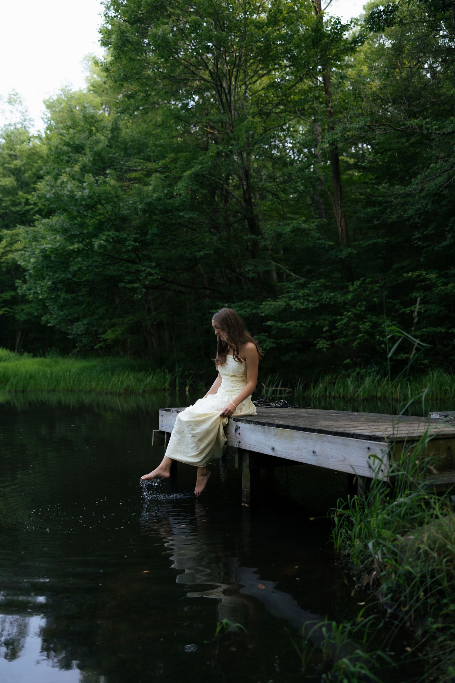 Young woman in cream dress sitting on wooden dock dipping feet in calm forest pond.