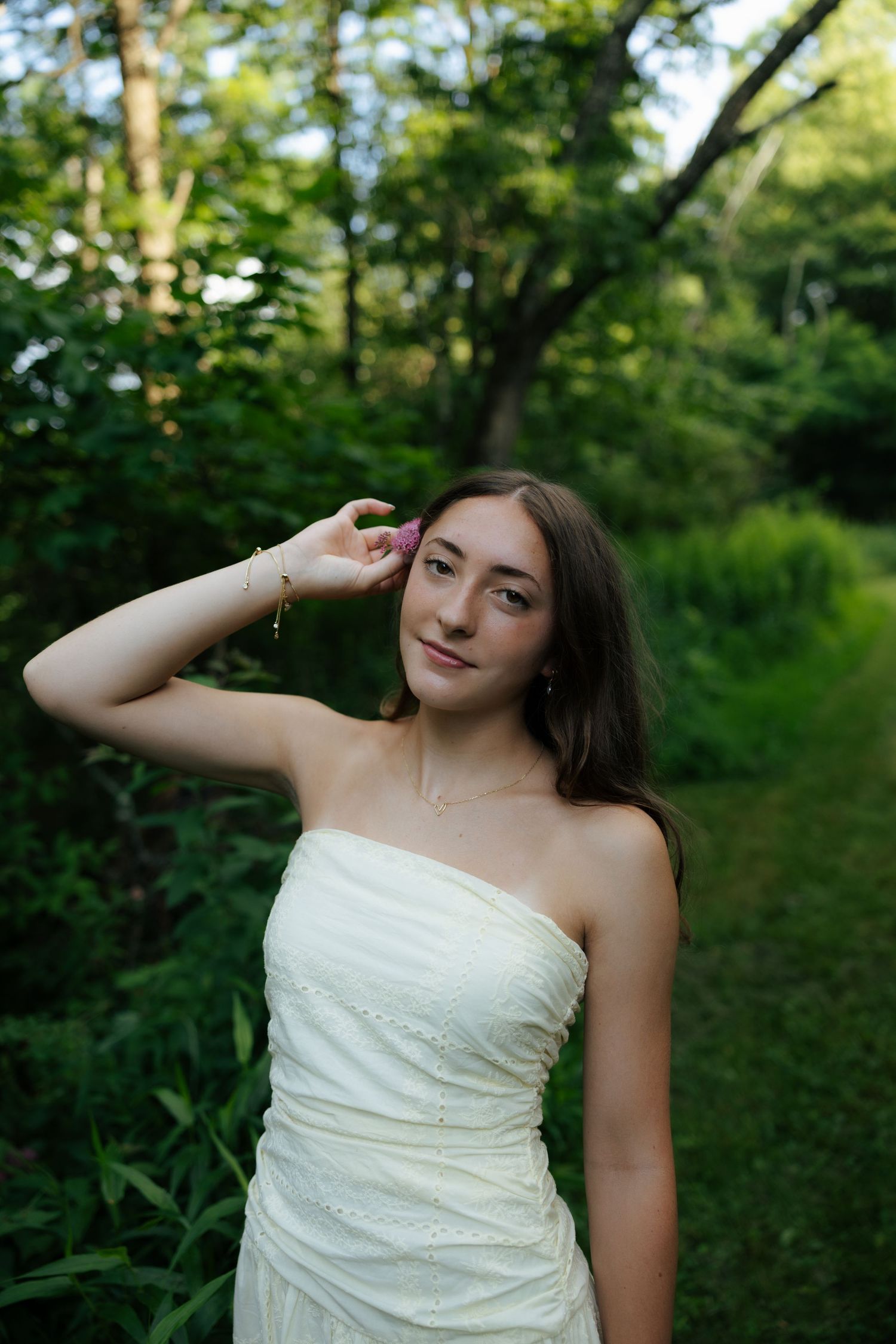 Young brunette woman in a strapless cream dress posing along a lush green garden pathway.