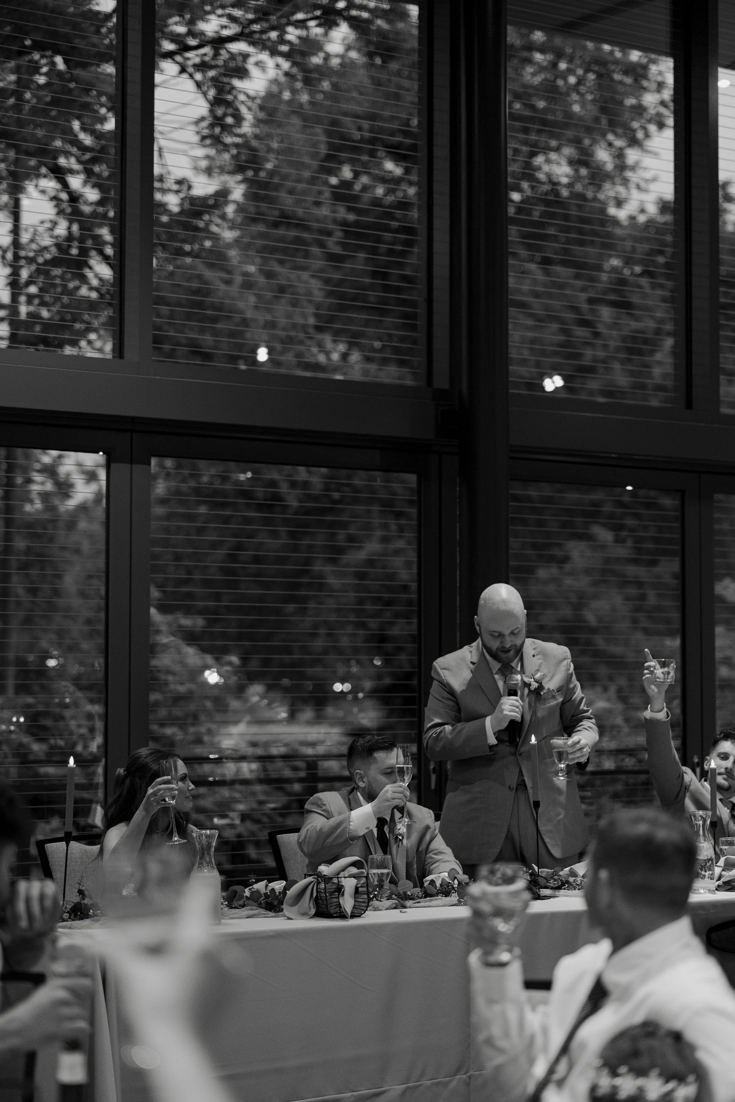 Black and white wedding reception toast with guests raising glasses as a man in suit speaks at the head table.