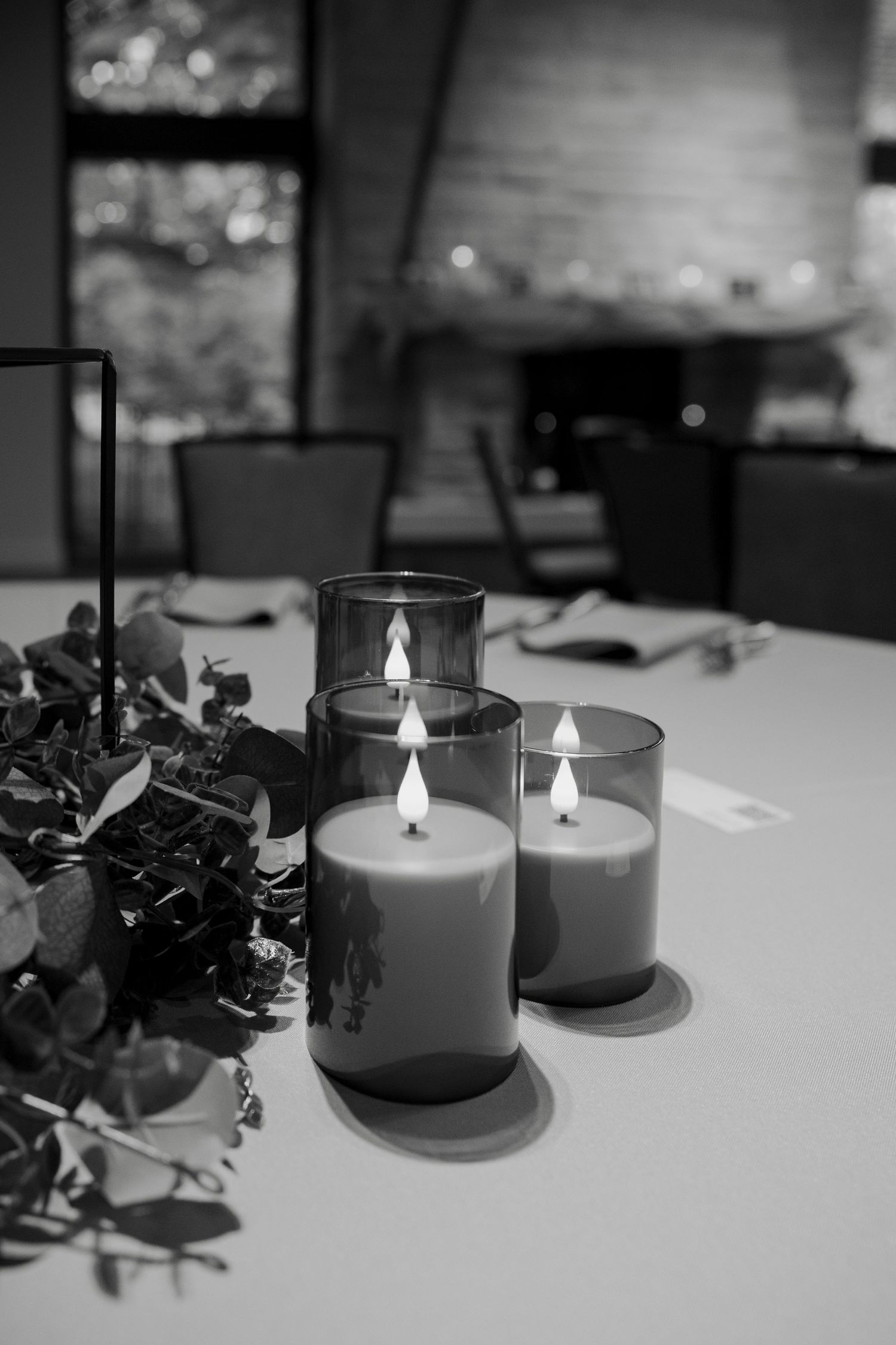 Black and white photo of three lit candles in glass holders on a table with greenery decor at an elegant event.